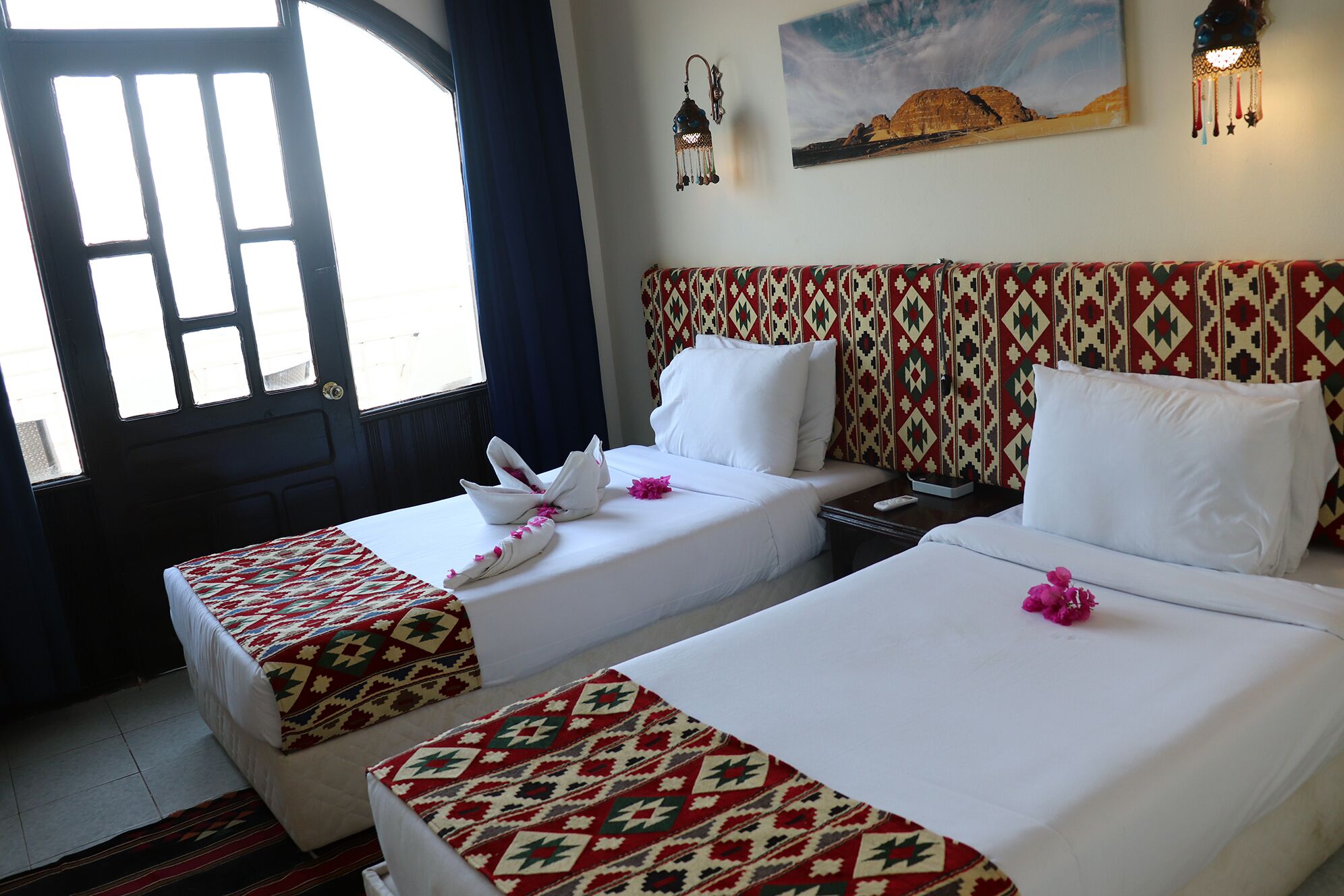 A hotel room with two beds, each with patterned bedspreads and white pillows. There is a window with curtains, a painting on the wall, and a wall-mounted lamp.
