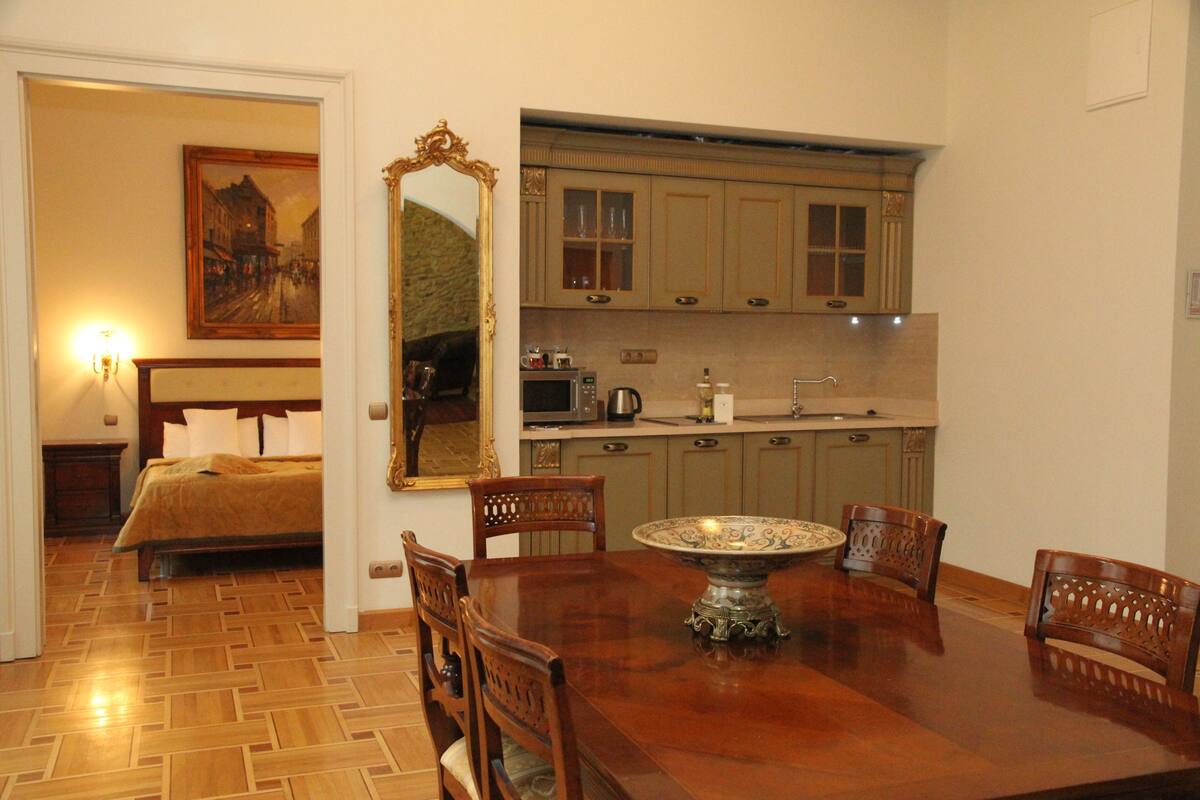 A hotel room with a wooden floor, a dining table with chairs, a kitchen area with cabinets, a bed, and a painting on the wall.
