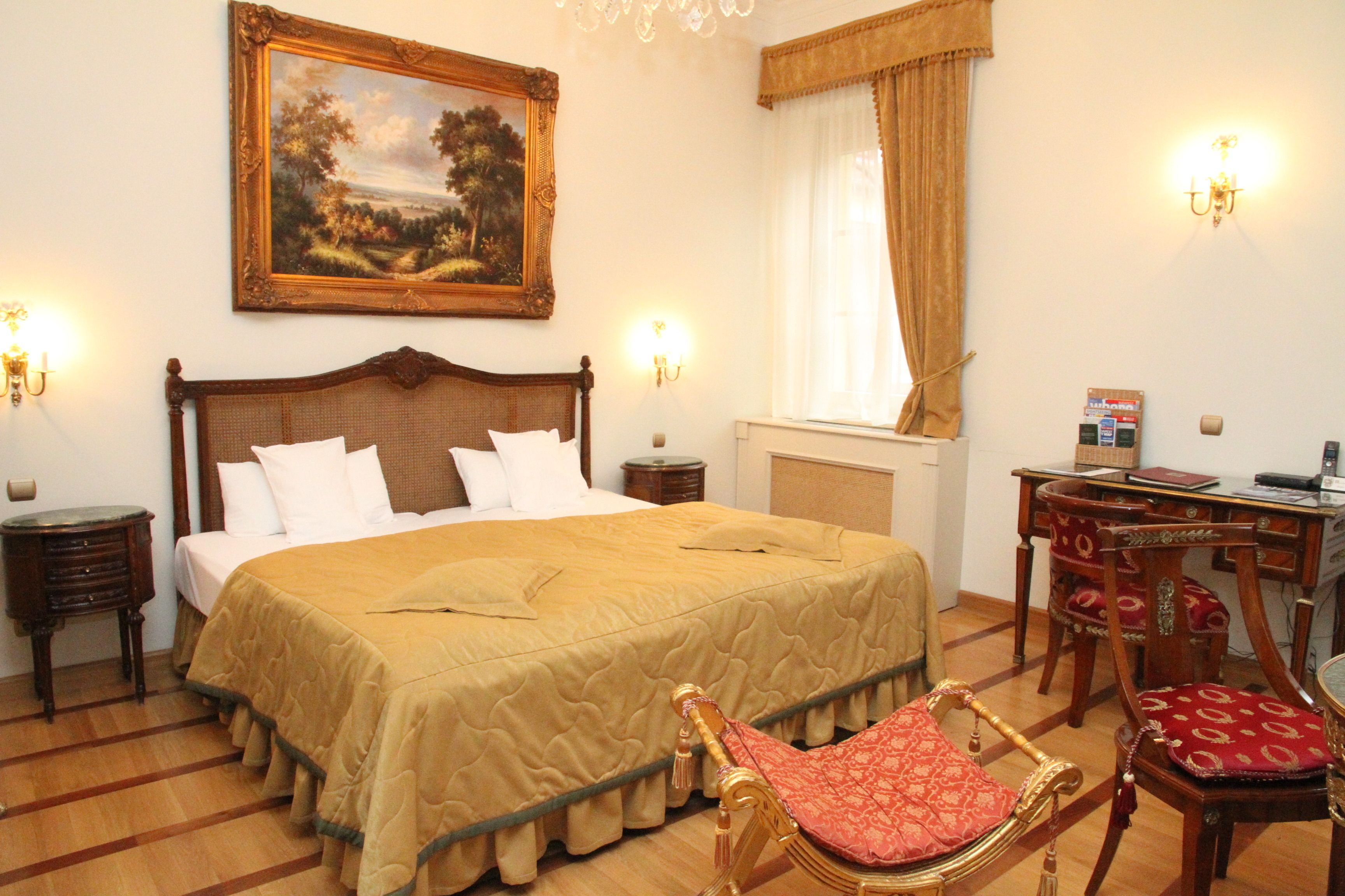 A hotel room with a large bed, a desk with a chair, a painting on the wall, and a chandelier.