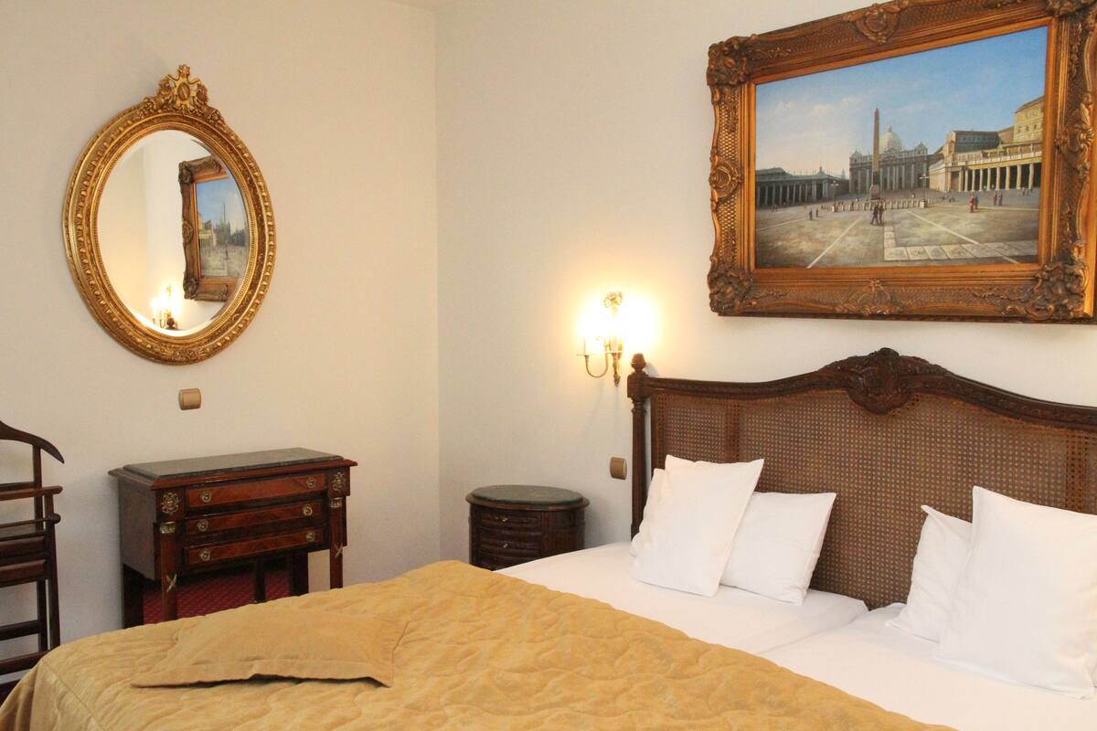 A hotel room with a large bed, a wooden headboard, a mirror, a dresser, and a painting of a cityscape on the wall.