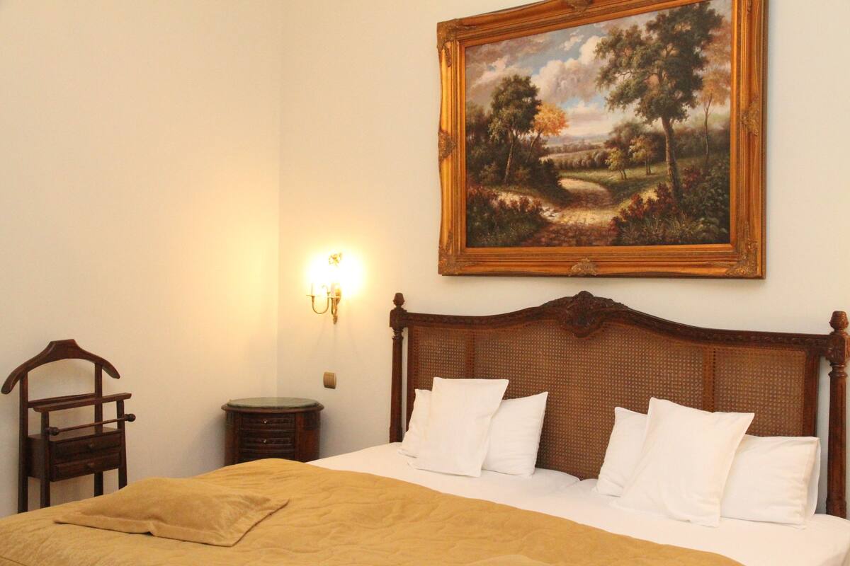 A neatly made bed with white linens and a brown blanket, a wooden headboard, a nightstand with a lamp, and a framed landscape painting on the wall.