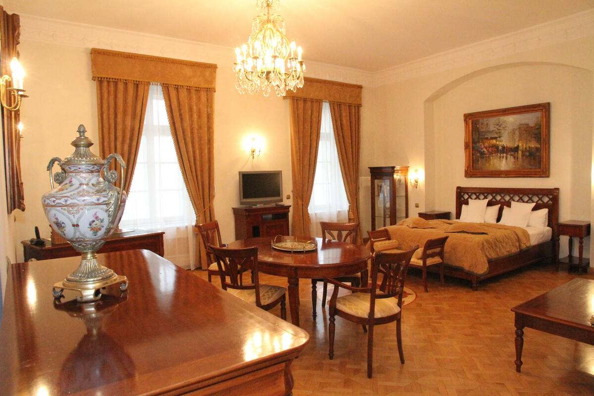 A hotel room with a wooden dining table, chairs, a chandelier, a bed with two pillows, a television, and a painting on the wall.
