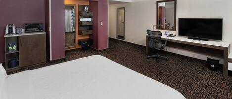 A hotel room with a bed, a desk with a TV, a chair, a minibar, and a bathroom with a shower curtain.