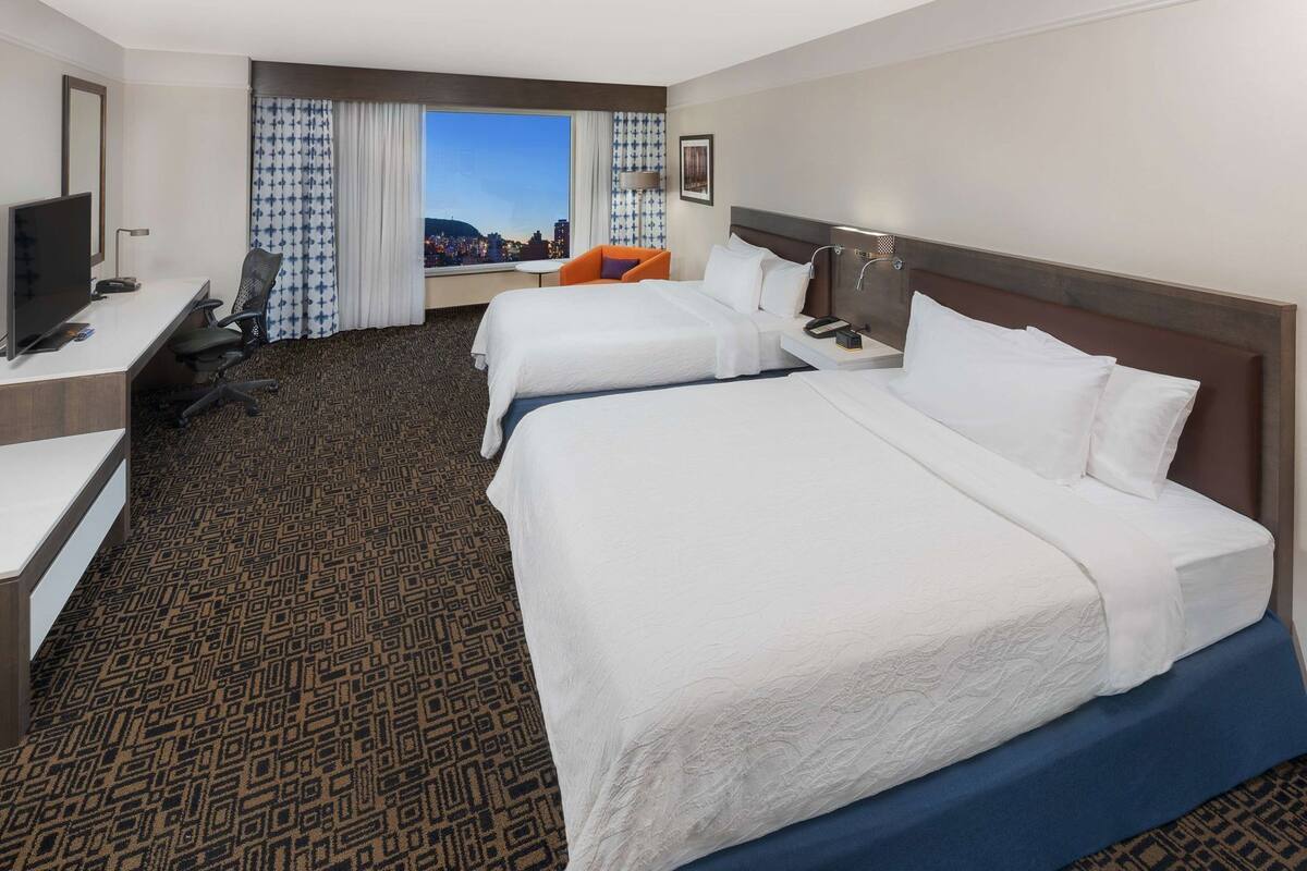 A hotel room with two beds, a desk with a chair, a TV, and a city view.