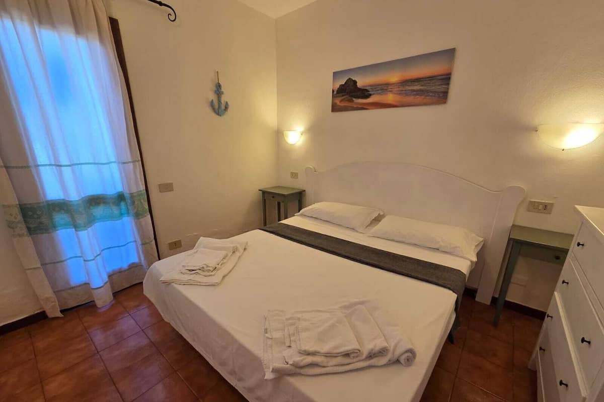 Un lit bien fait, avec du linge de lit et des serviettes blanches, un tableau représentant un coucher de soleil accroché au mur et une petite table de chevet avec une lampe.