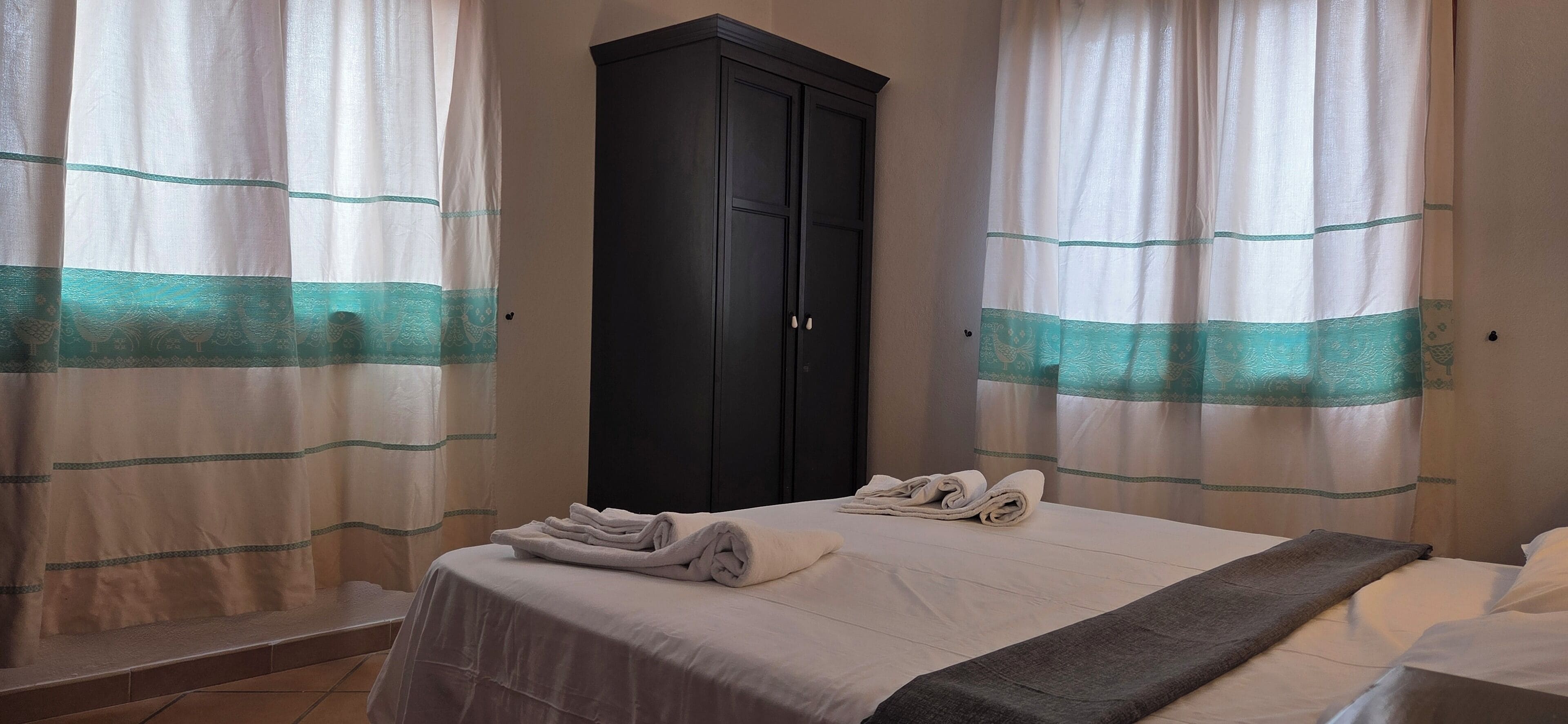 A hotel room with a bed, a dark wardrobe, and curtains with a teal pattern.