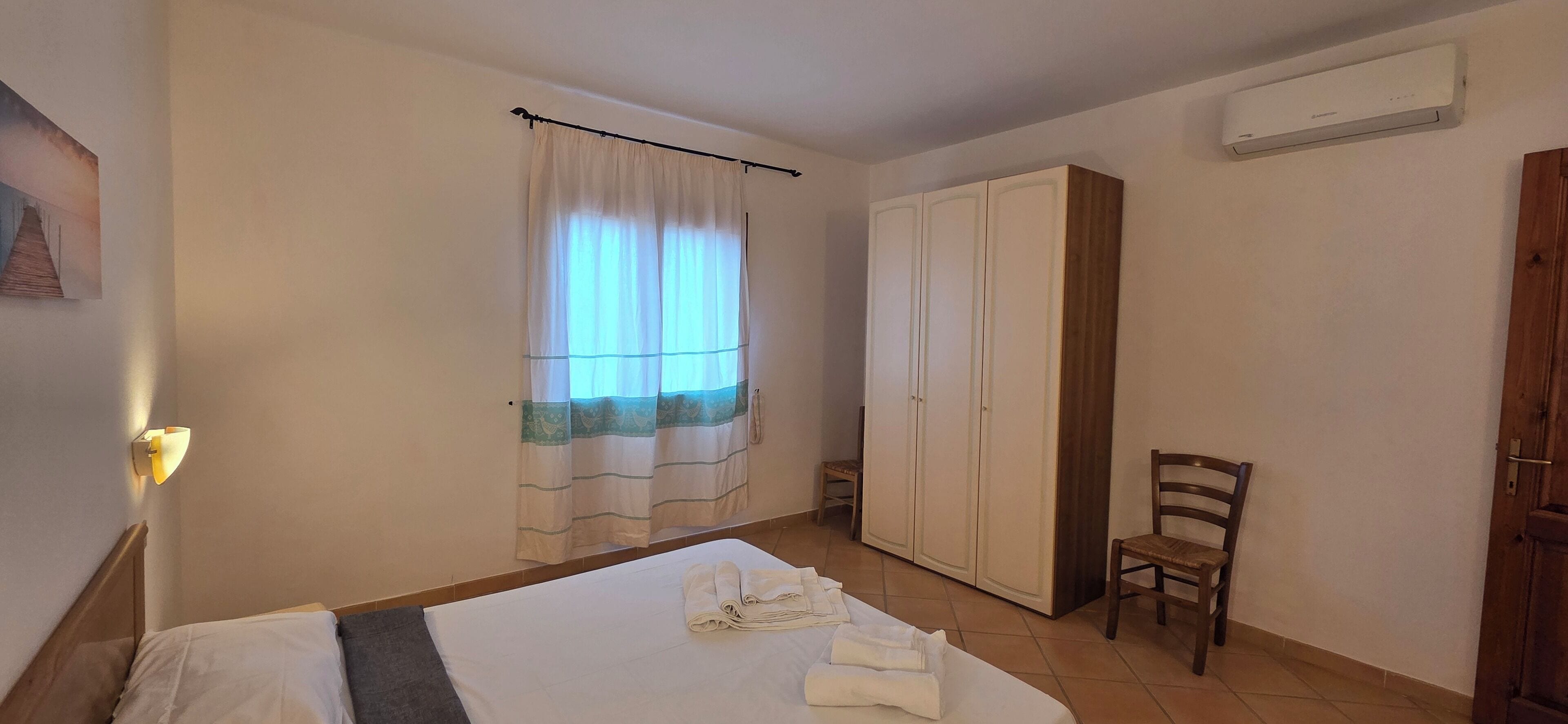 A hotel room with a bed, a wardrobe, a chair, a window with curtains, and an air conditioning unit.