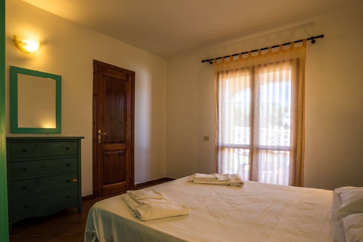 Une chambre à coucher avec un lit, une commode, une porte et une fenêtre avec des rideaux.
