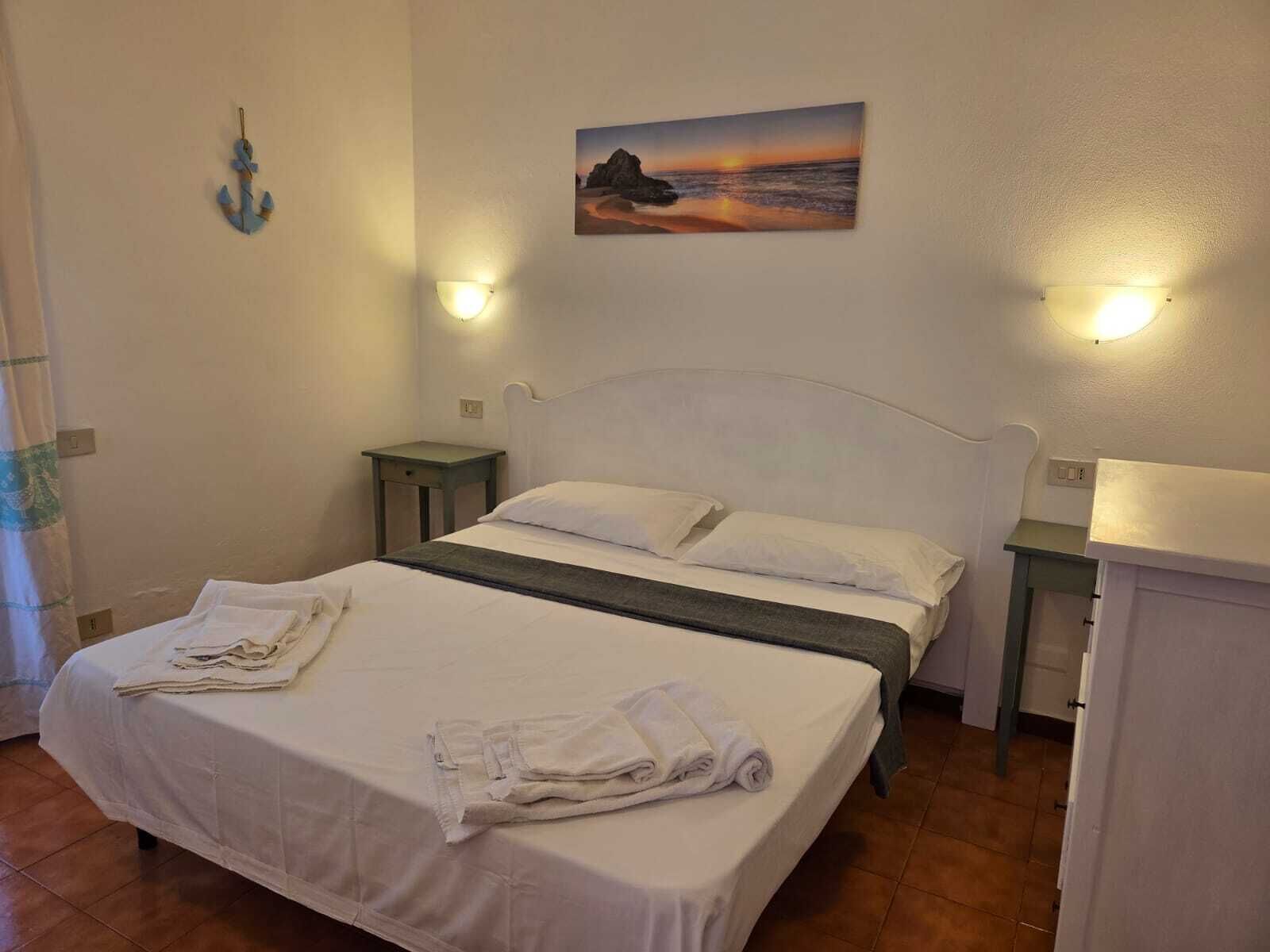 A neatly made bed with white linens and towels, two bedside tables, a wall-mounted painting of a sunset, and a small decorative anchor.