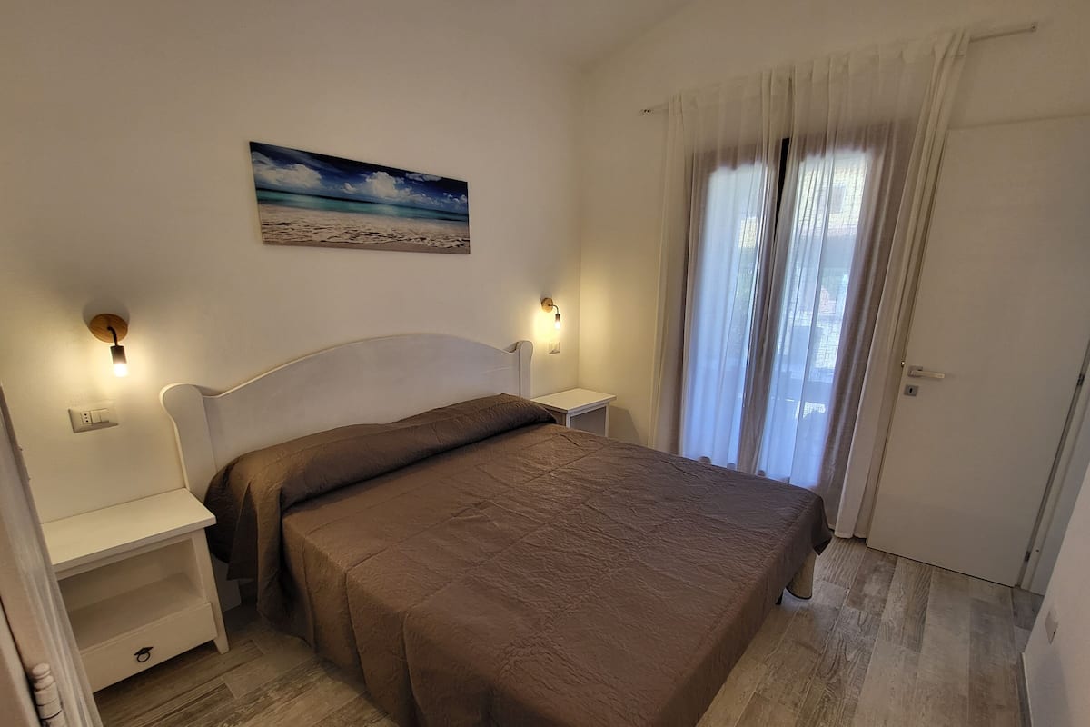Une chambre à coucher avec un lit, des tables de chevet, une commode, un tableau et une fenêtre avec des rideaux.