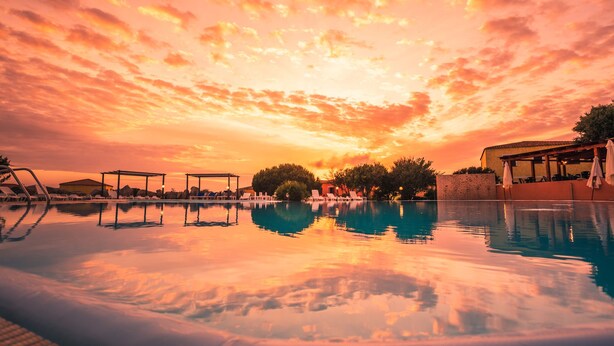 A swimming pool with lounge chairs and umbrellas, surrounded by buildings and trees, reflecting a sunset sky. - RESIDENCE LU LAMONI COSTA NORD (Aglientu)