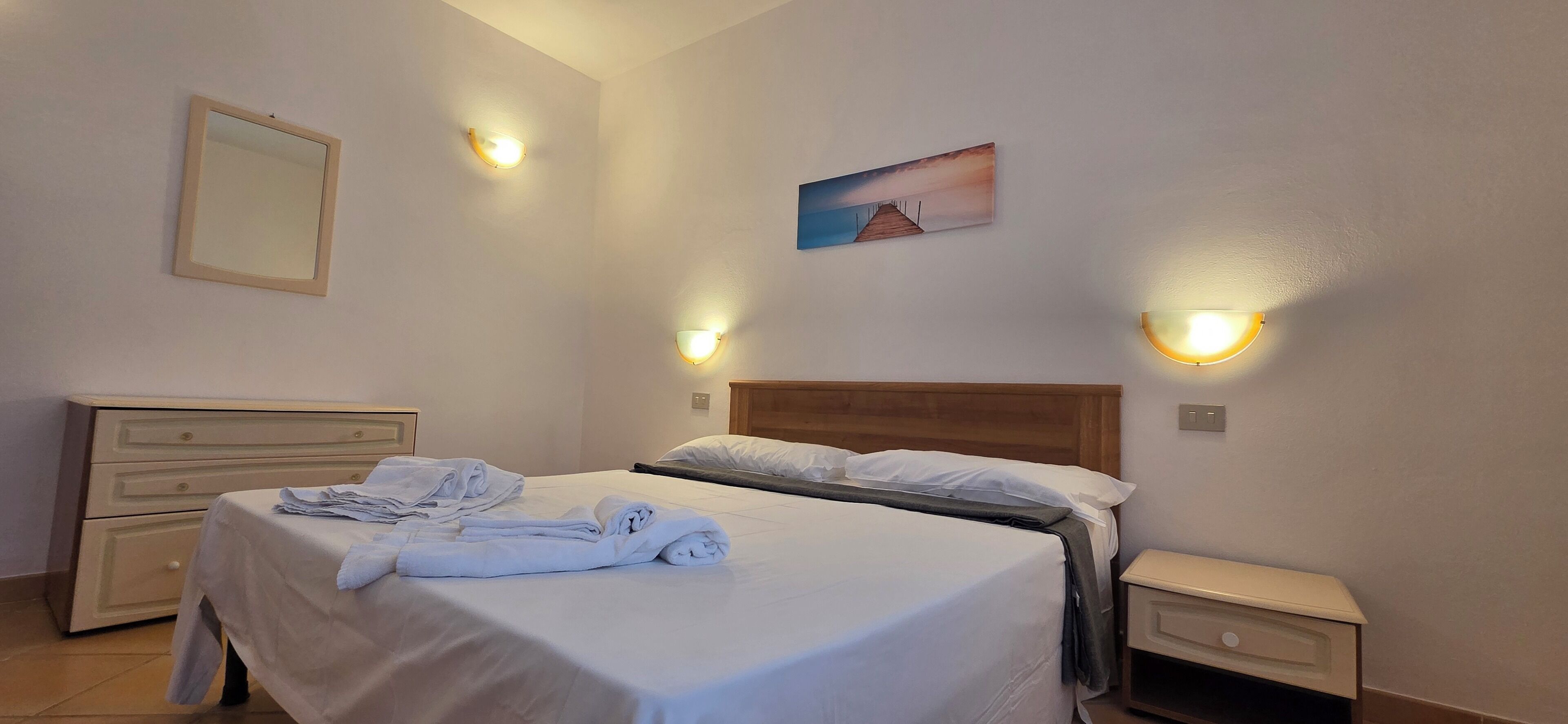A neatly made bed with white linens, a bedside table, a nightstand, a wall-mounted mirror, and two wall sconces.