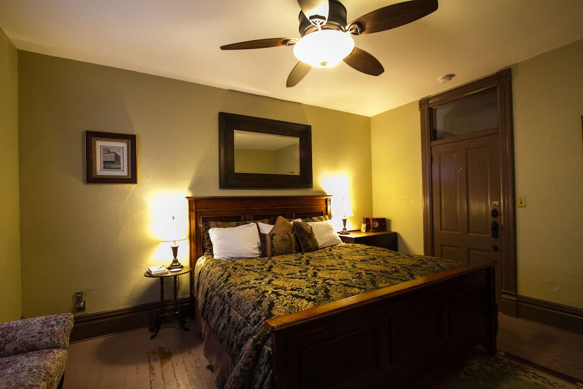 A bedroom with a bed, a ceiling fan, a nightstand, a lamp, a framed picture, and a door.