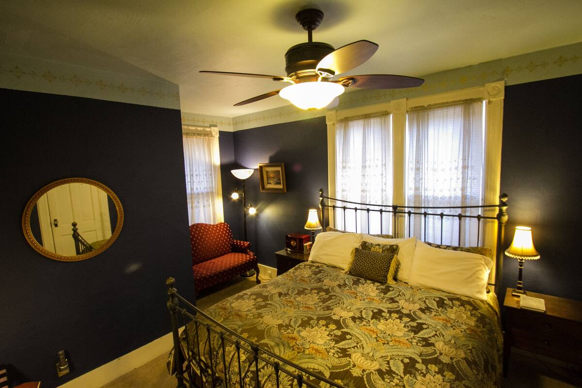 A bedroom with a bed, a chair, a nightstand, a lamp, a ceiling fan, and a window with curtains.