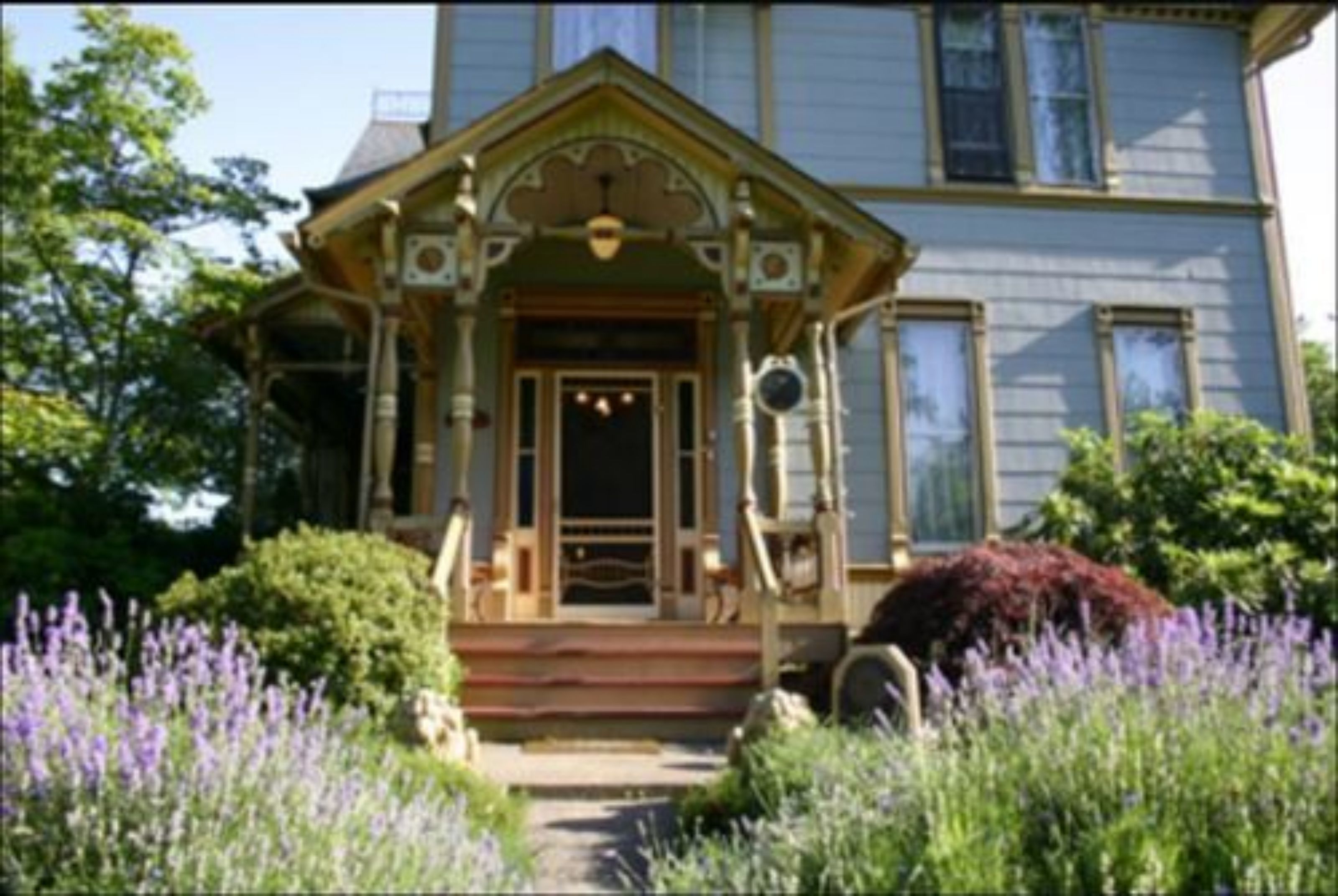 A Victorian-style house with a front porch, steps, and a garden with purple flowers.