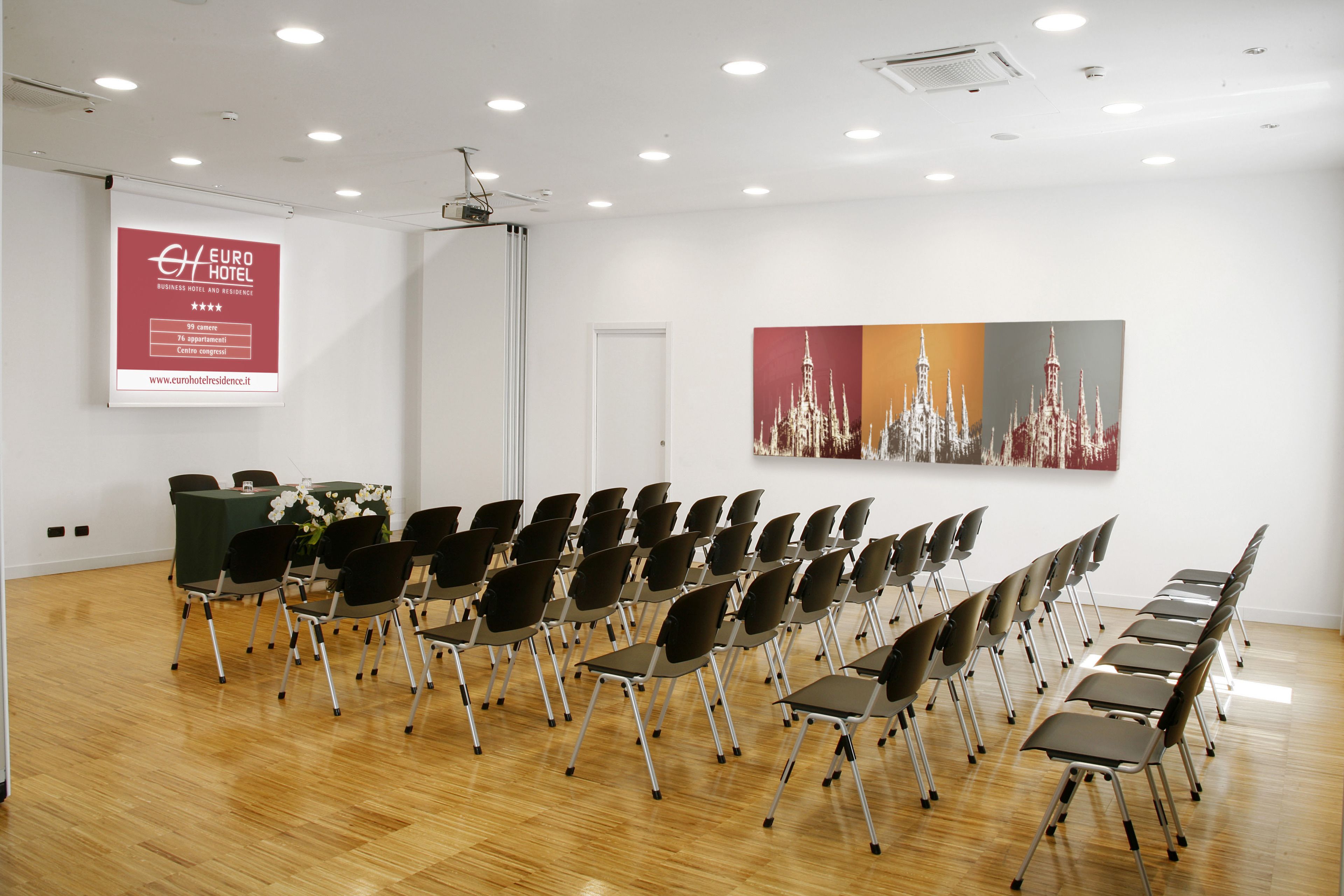 A conference room with rows of chairs, a projector screen, and a large painting on the wall.
