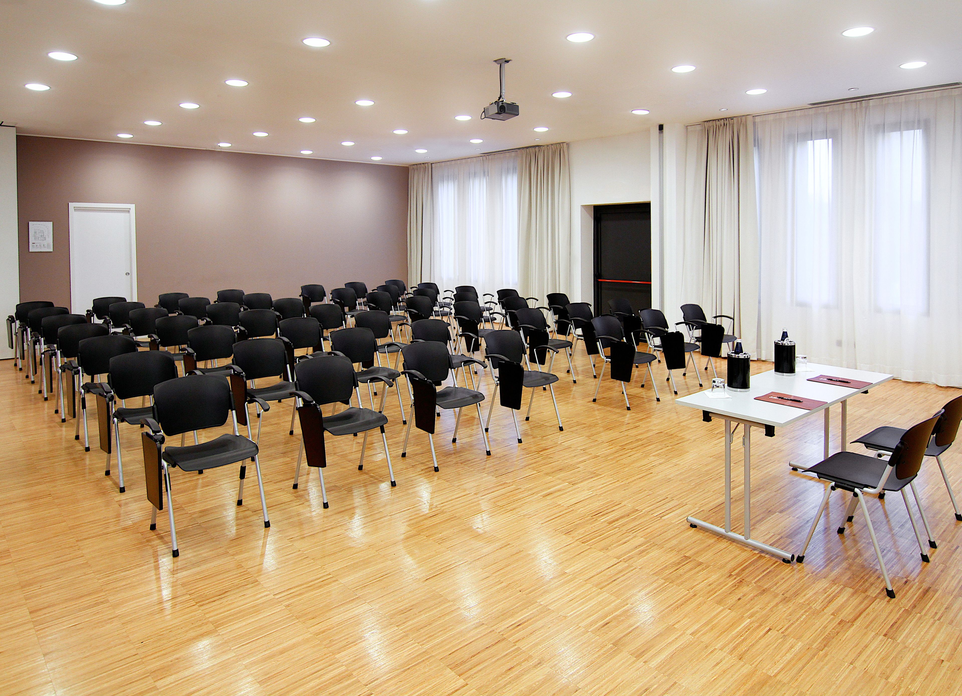 Una sala conferenze con file di sedie, un tavolo bianco con un proiettore e un'ampia finestra con tende.