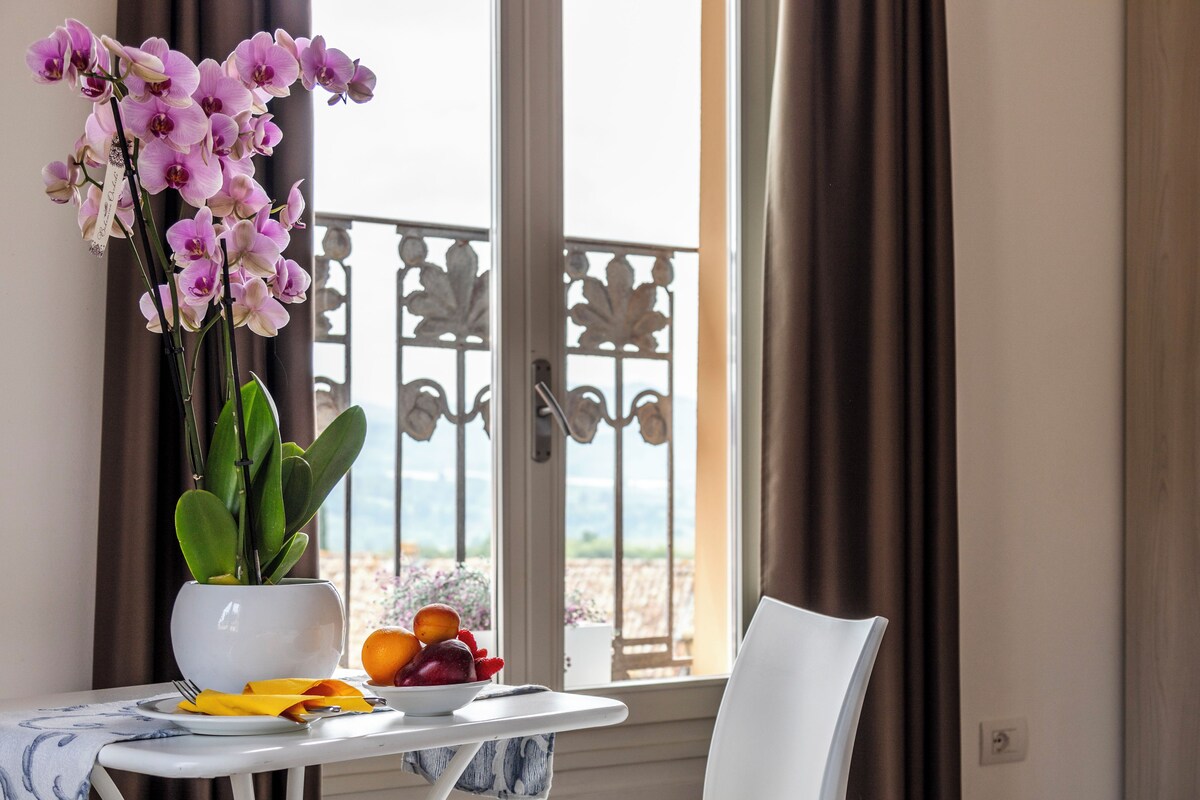 Una mesa con un jarrón de flores, un frutero y una silla.