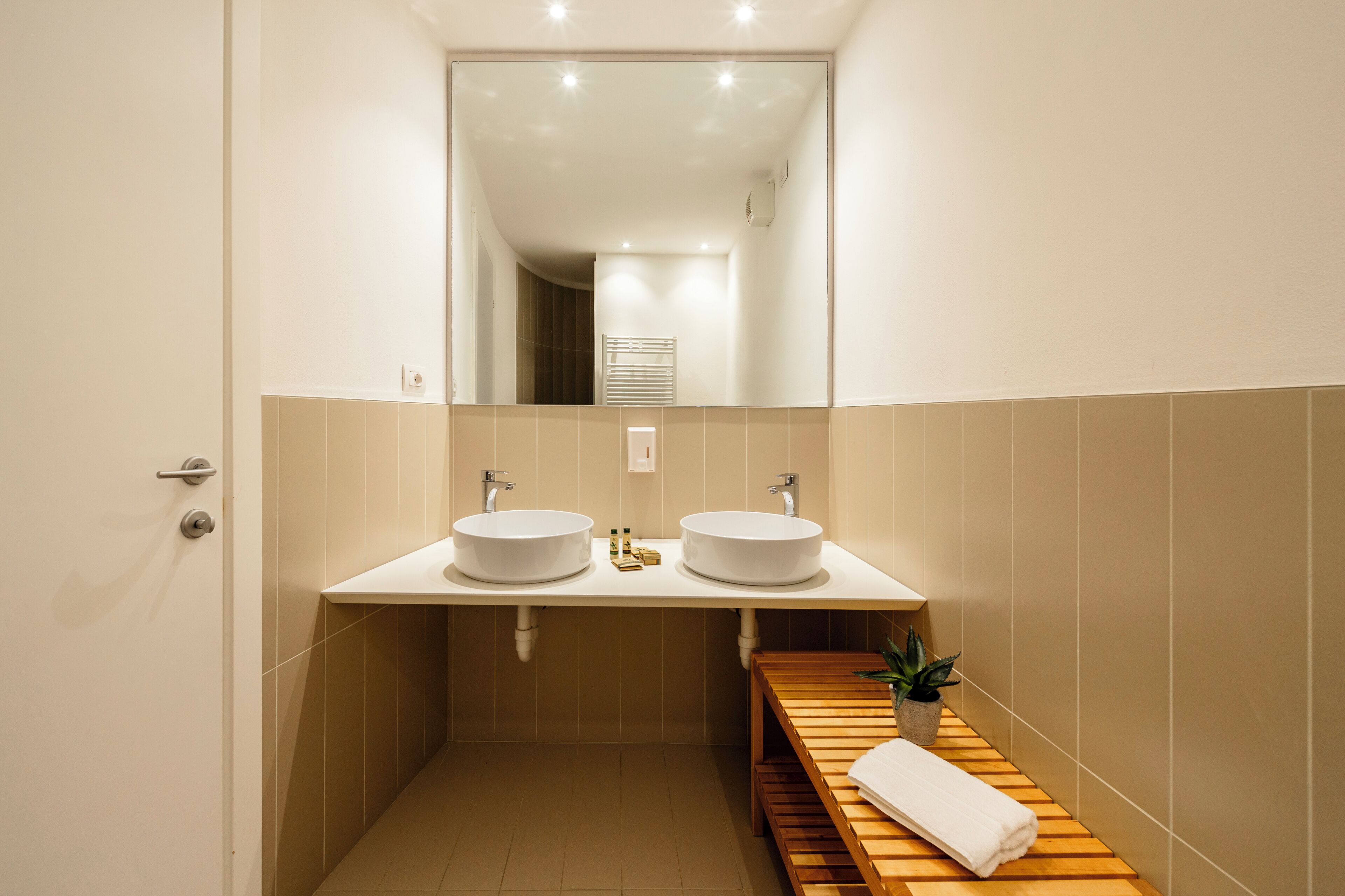 A modern bathroom with two sinks, a mirror, and a wooden bench with a towel.