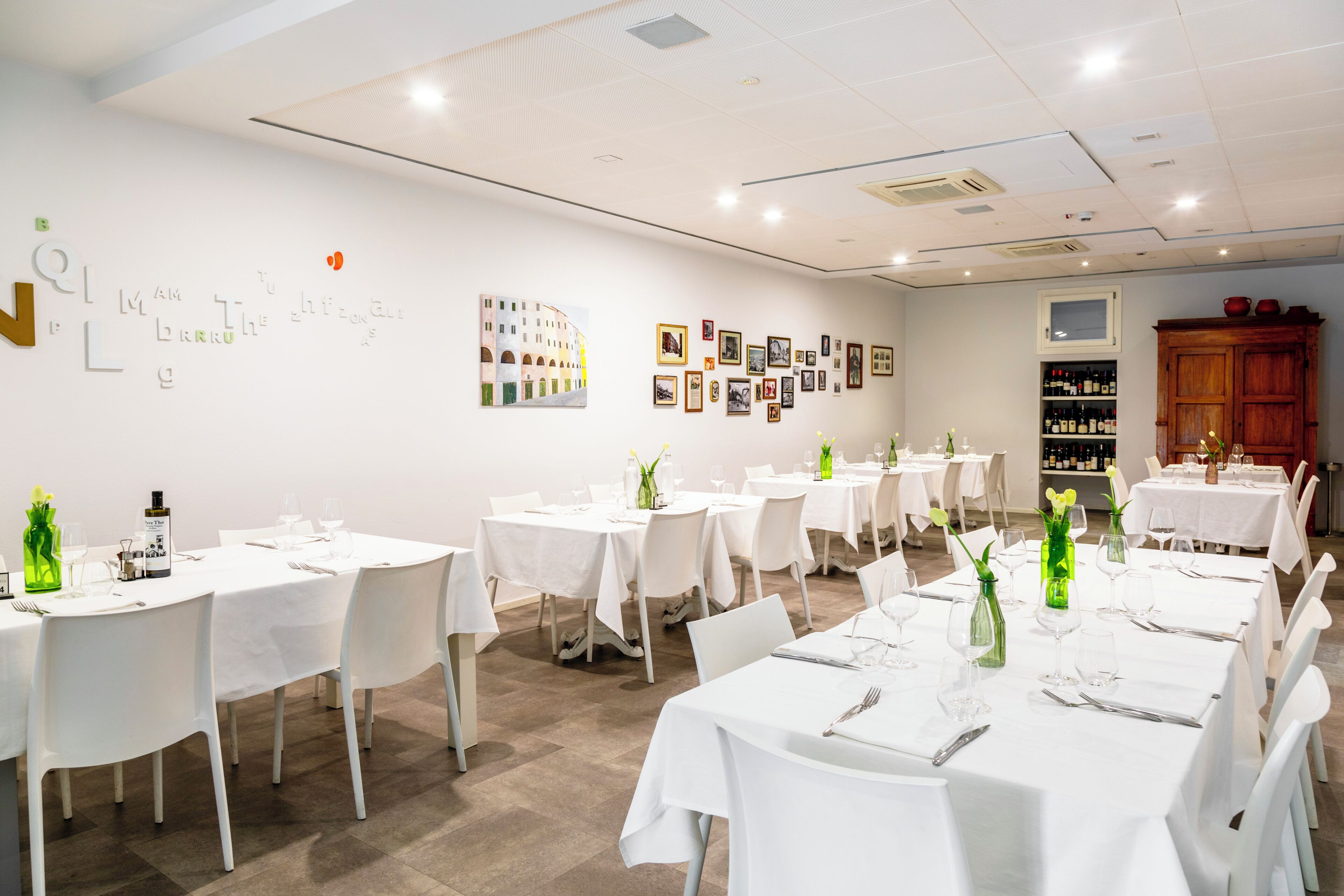 A dining area with white tables and chairs, arranged in rows, with artwork on the walls and a wine rack in the background.