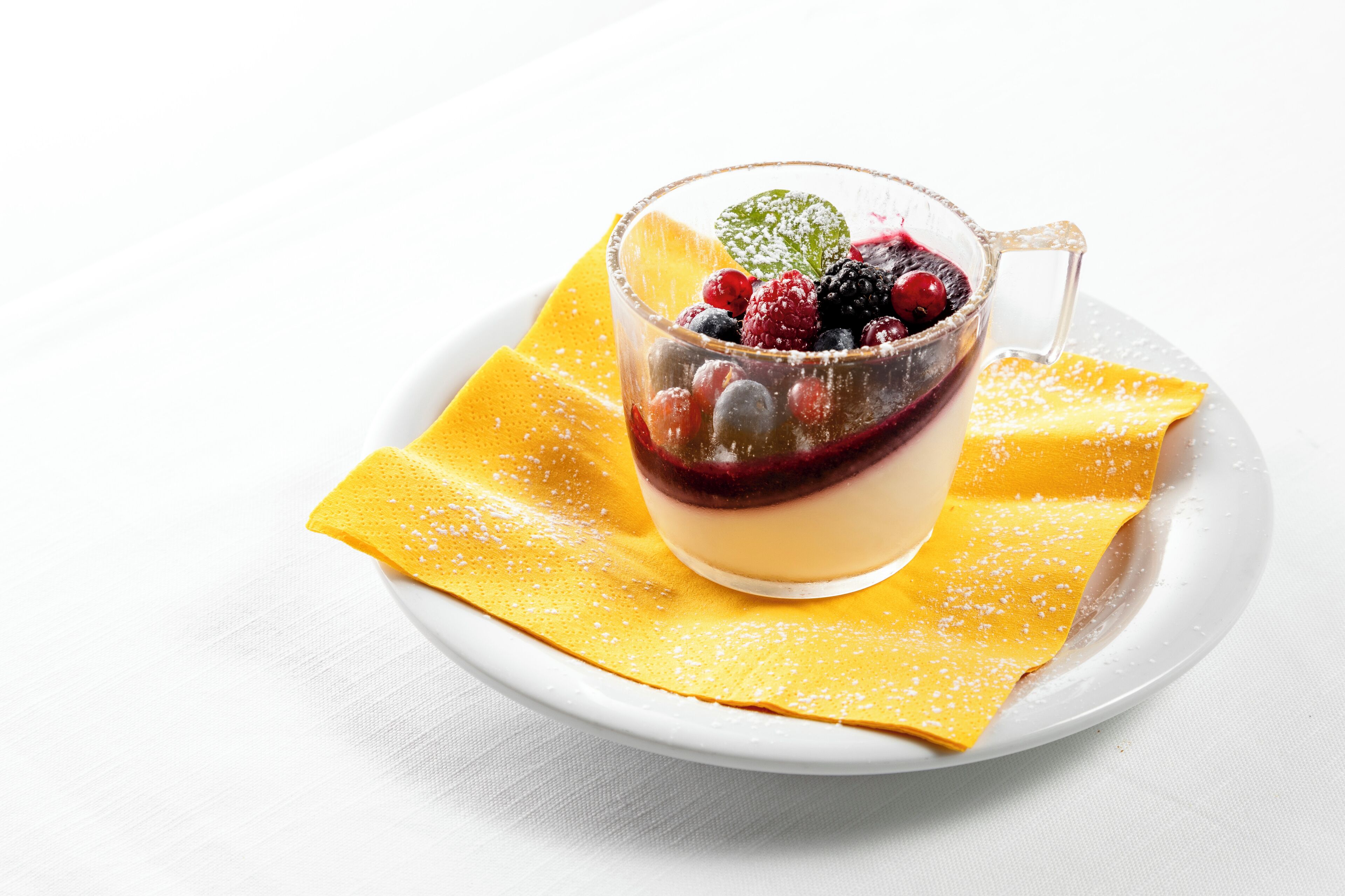 A dessert with berries and a mint leaf on a clear glass, served on a yellow napkin and a white plate.