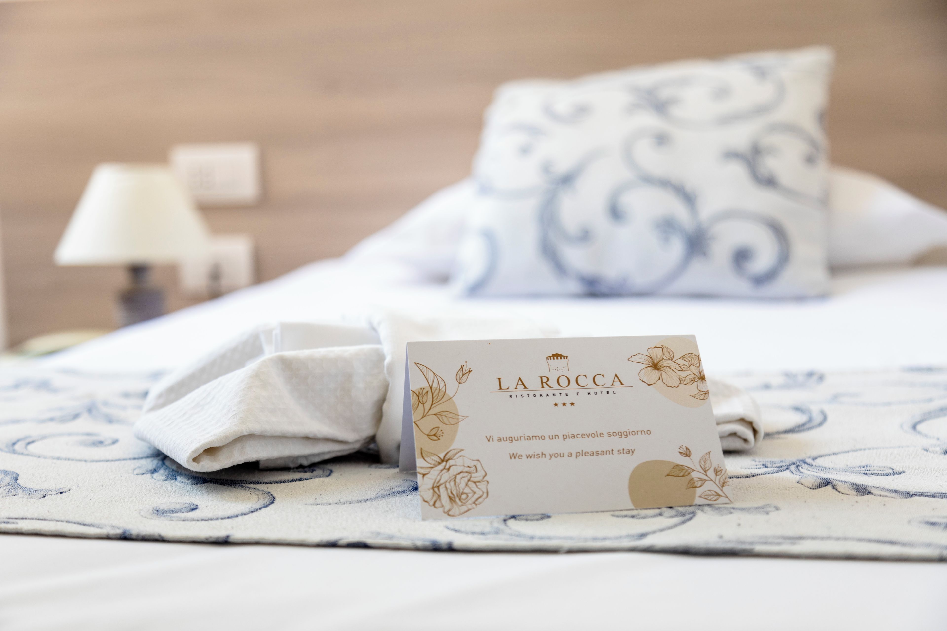 A neatly made bed with a white and blue patterned bedspread, a folded white towel, and a card from La Rocca Ristorante e Hotel.
