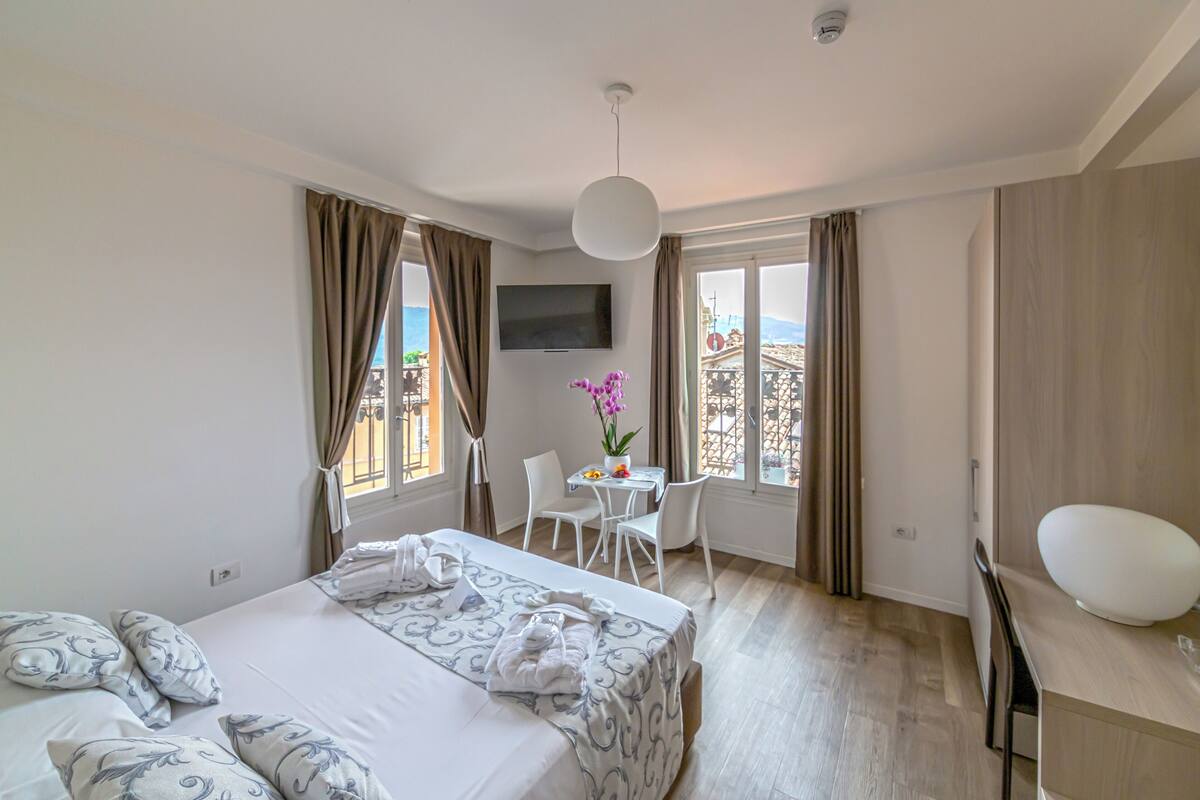 Habitación de hotel con cama, escritorio, silla, televisor y balcón con cortinas.