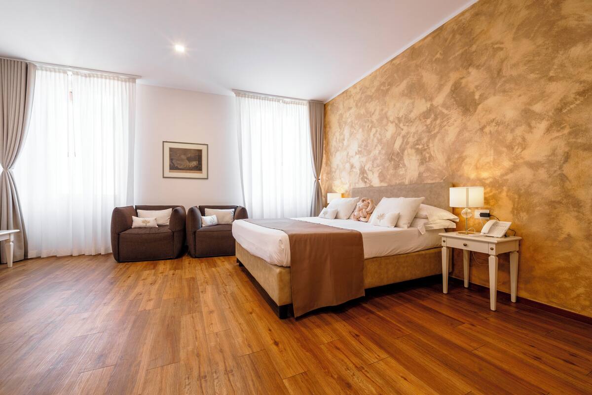 Habitación de hotel con cama, dos sillones, mesita de noche con lámpara y un cuadro en la pared.