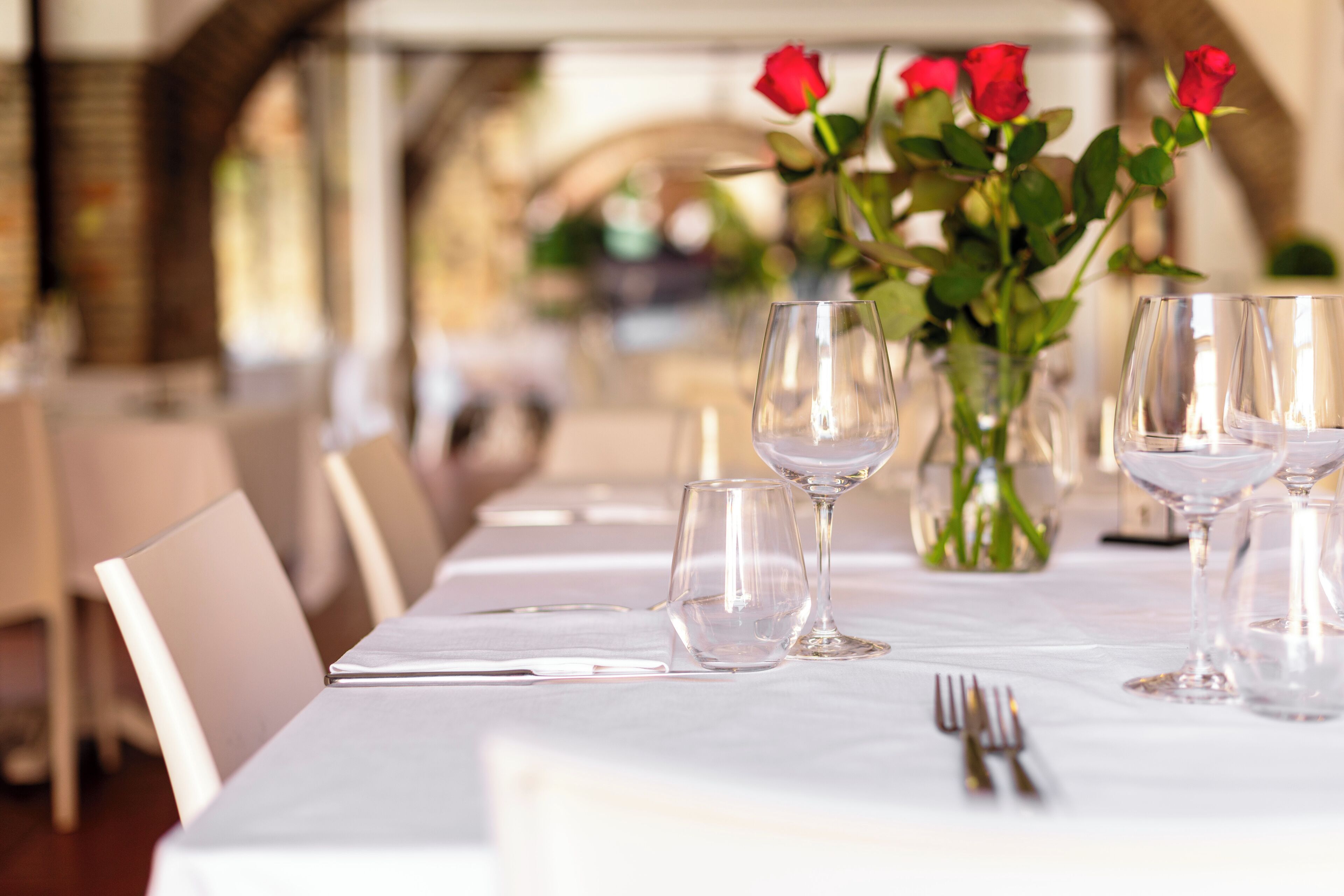 A restaurant table set with wine glasses, a vase with roses, and a white tablecloth.