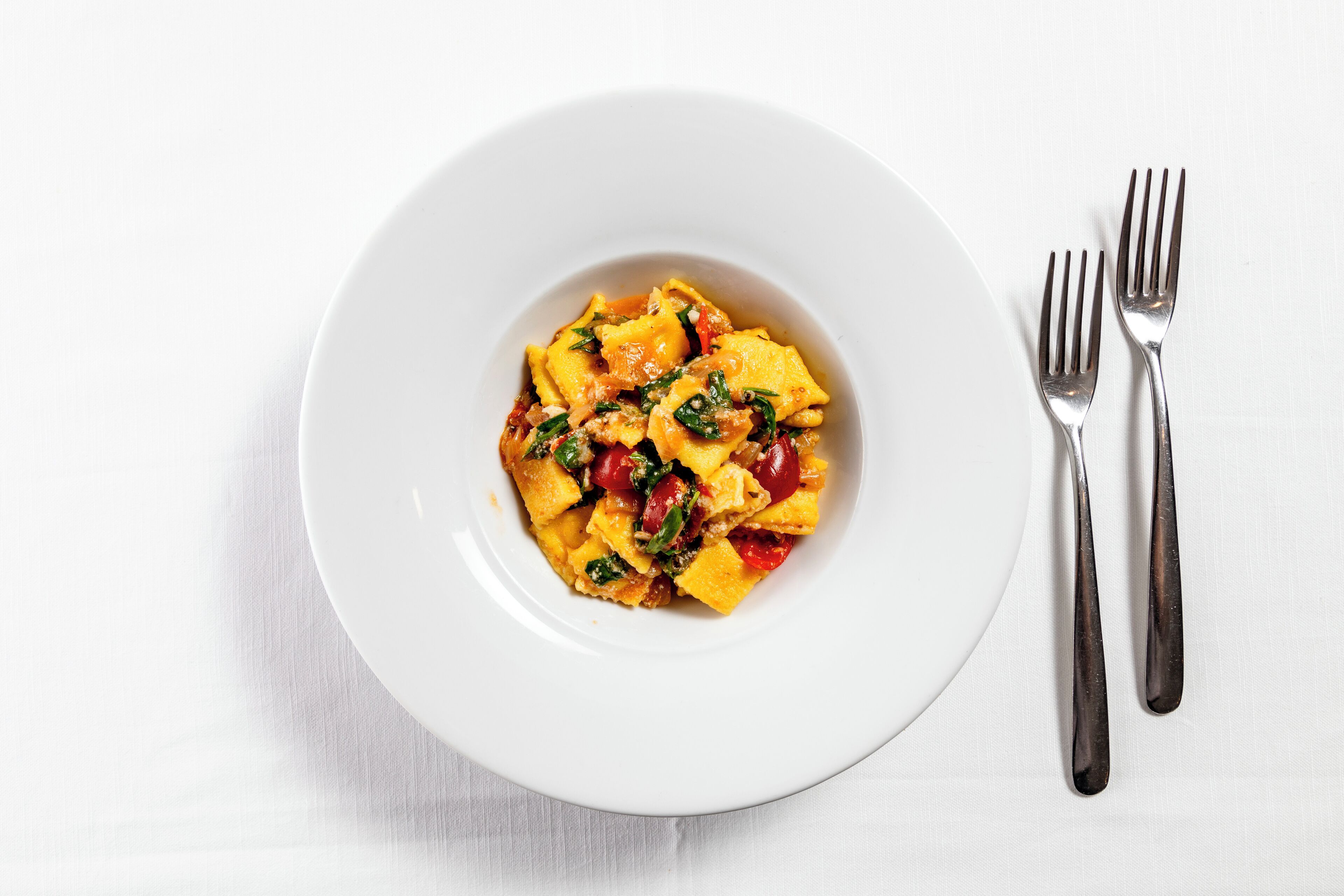 A white plate with a pasta dish and two forks on a white tablecloth.