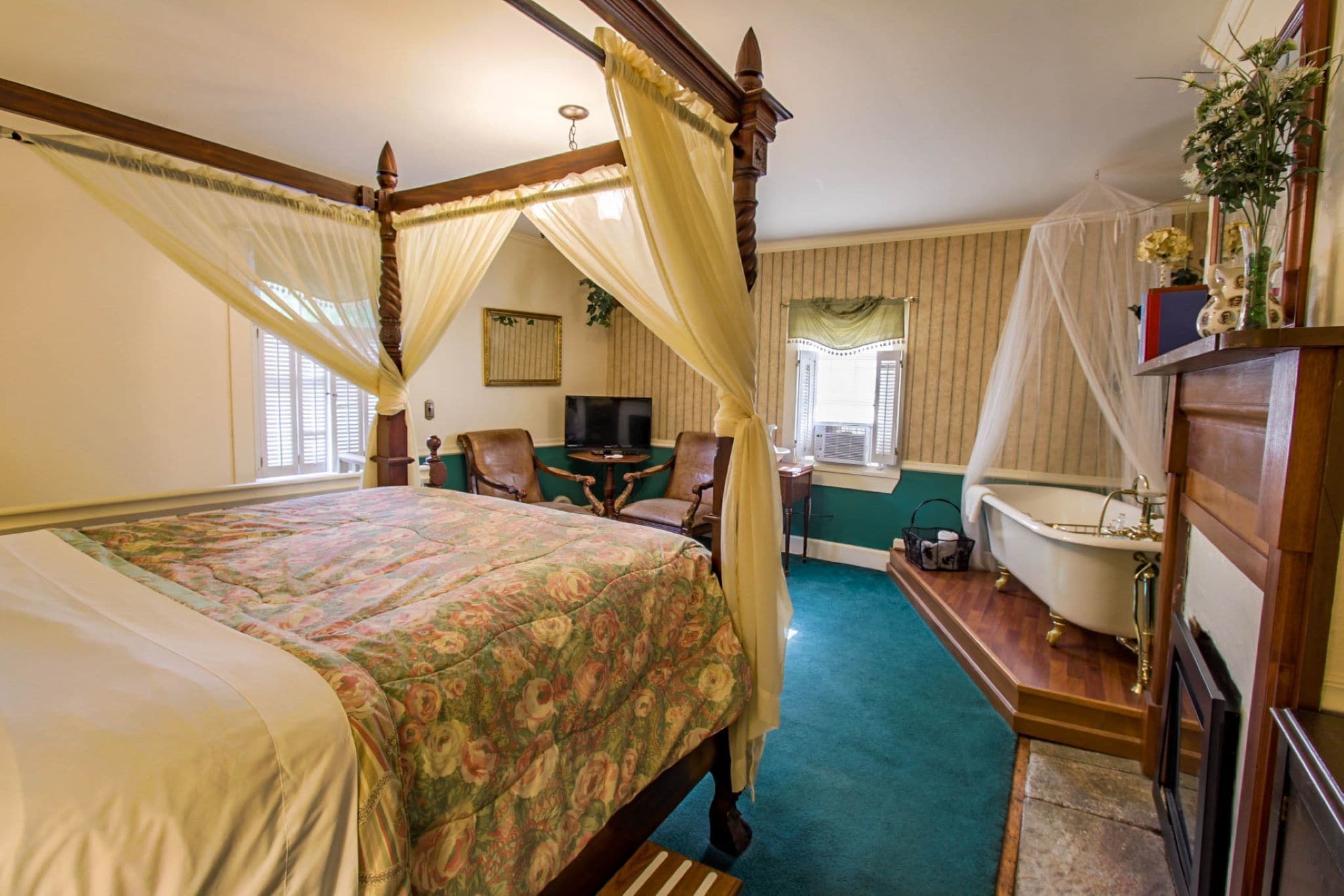 A bedroom with a four-poster bed, a sitting area with a table and chairs, a bathtub, and a television.