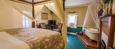 A bedroom with a four-poster bed, a sitting area with a table and chairs, a bathtub, and a television.