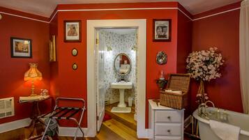 A room with red walls, a wooden floor, a white dresser, a white vanity, a white bathtub, a red rug, a mirror, and a door leading to another room.