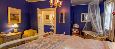 A bedroom with a large bed, a bedside table with a lamp, a chair, a vanity with a mirror, and a window with a curtain.