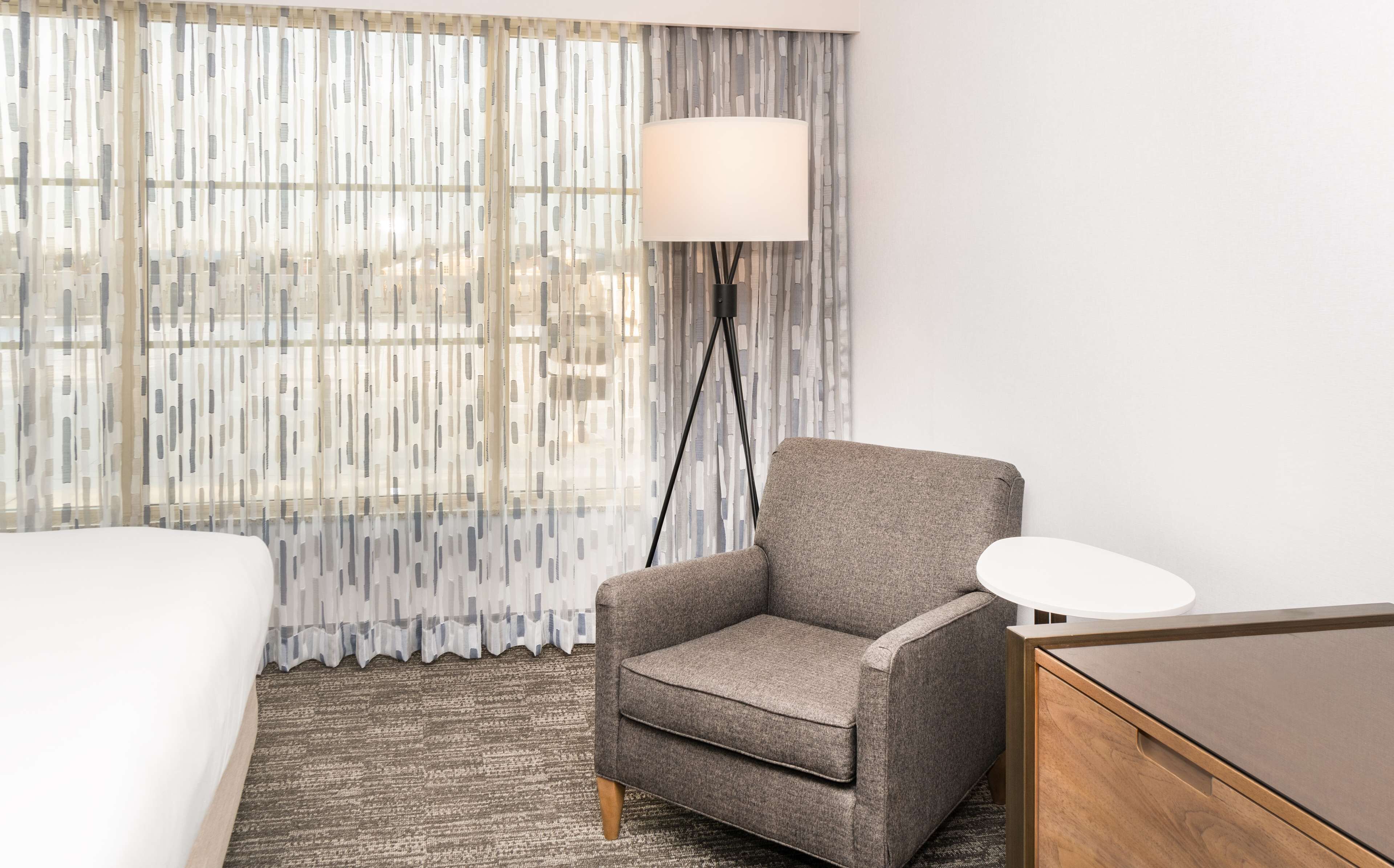 A hotel room with a grey armchair, a white bedside table, a lamp, and a bed.