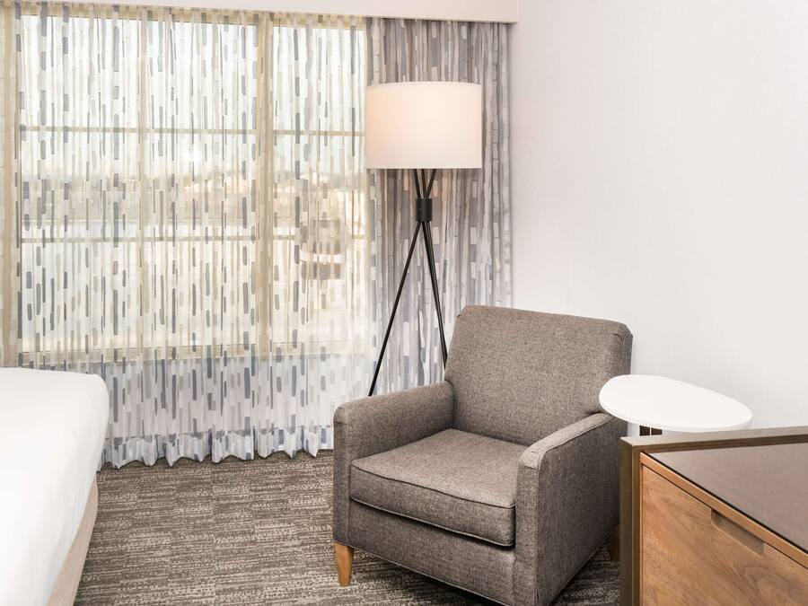 A hotel room with a grey armchair, a white bedside table, a lamp, and a bed.