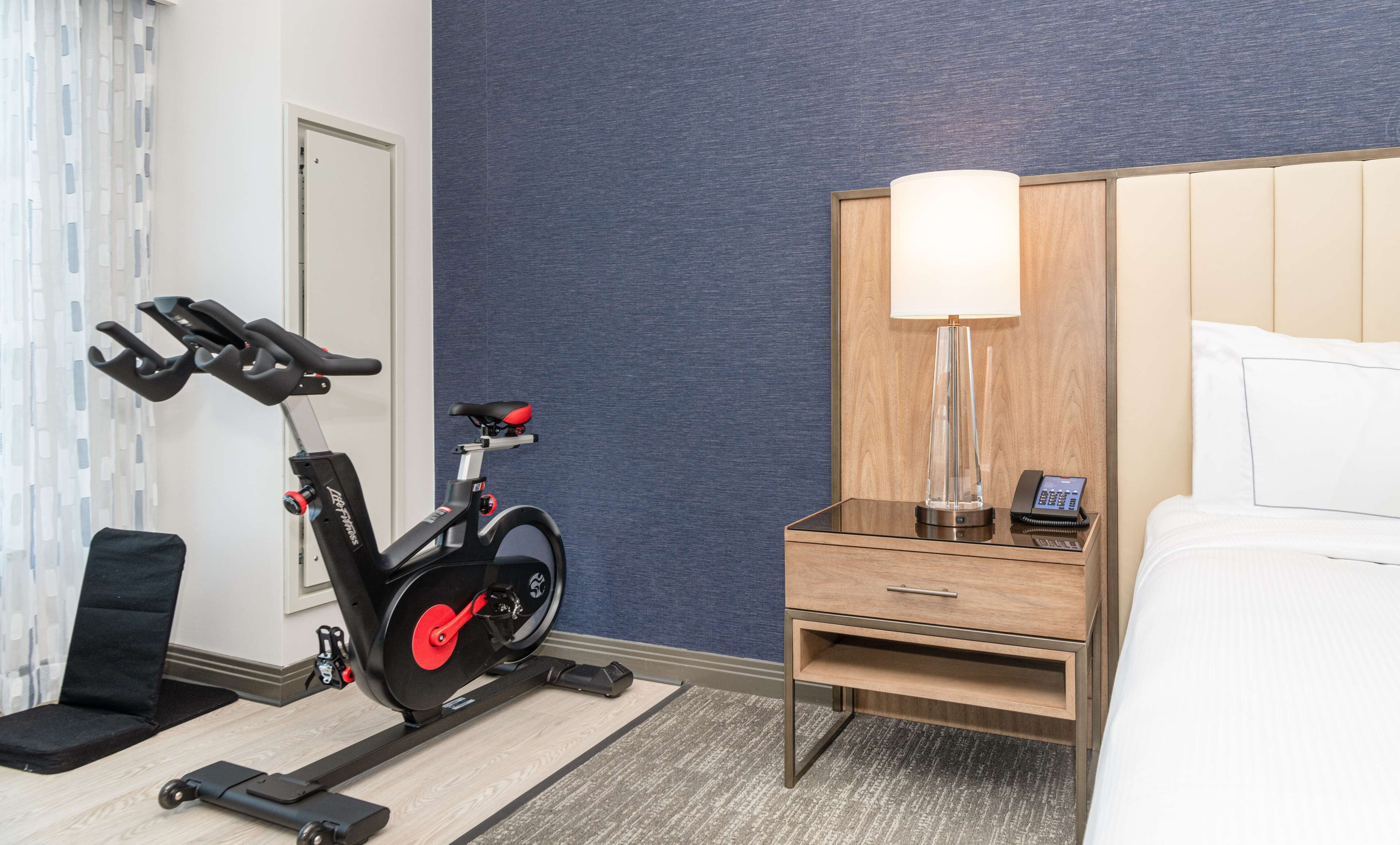 A hotel room with a stationary bike, a bedside table with a lamp, and a bed.
