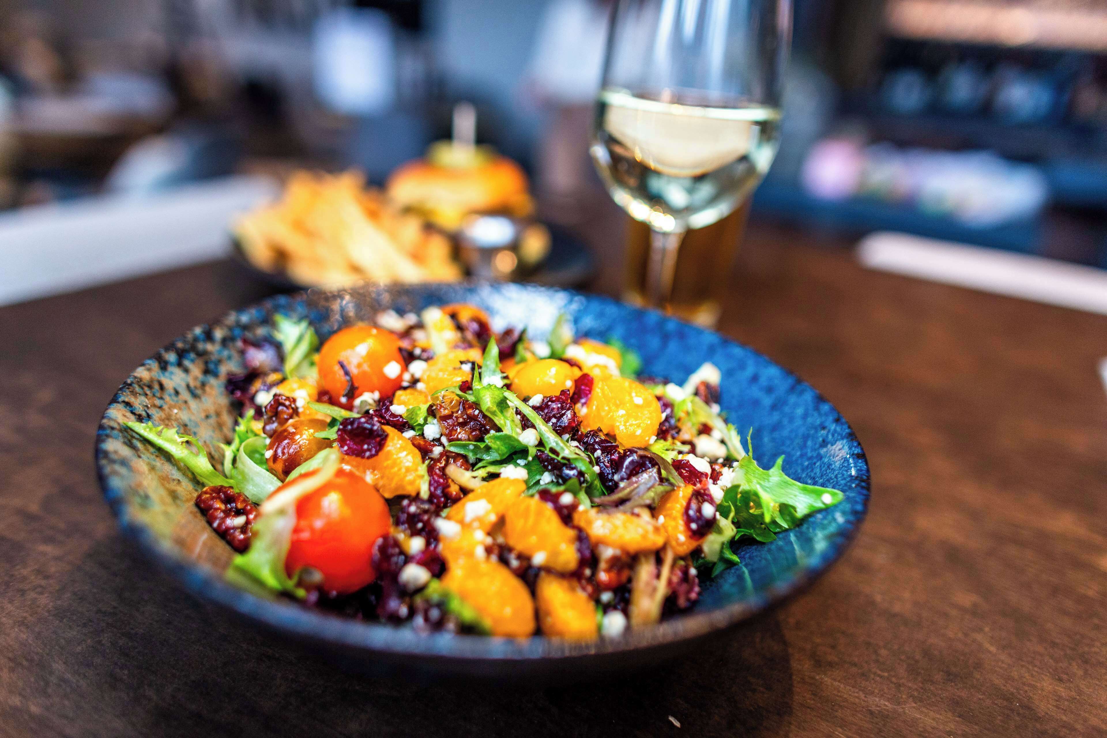 A plate of salad with various vegetables and fruits, a glass of white wine, and a blurred background suggesting a dining setting.