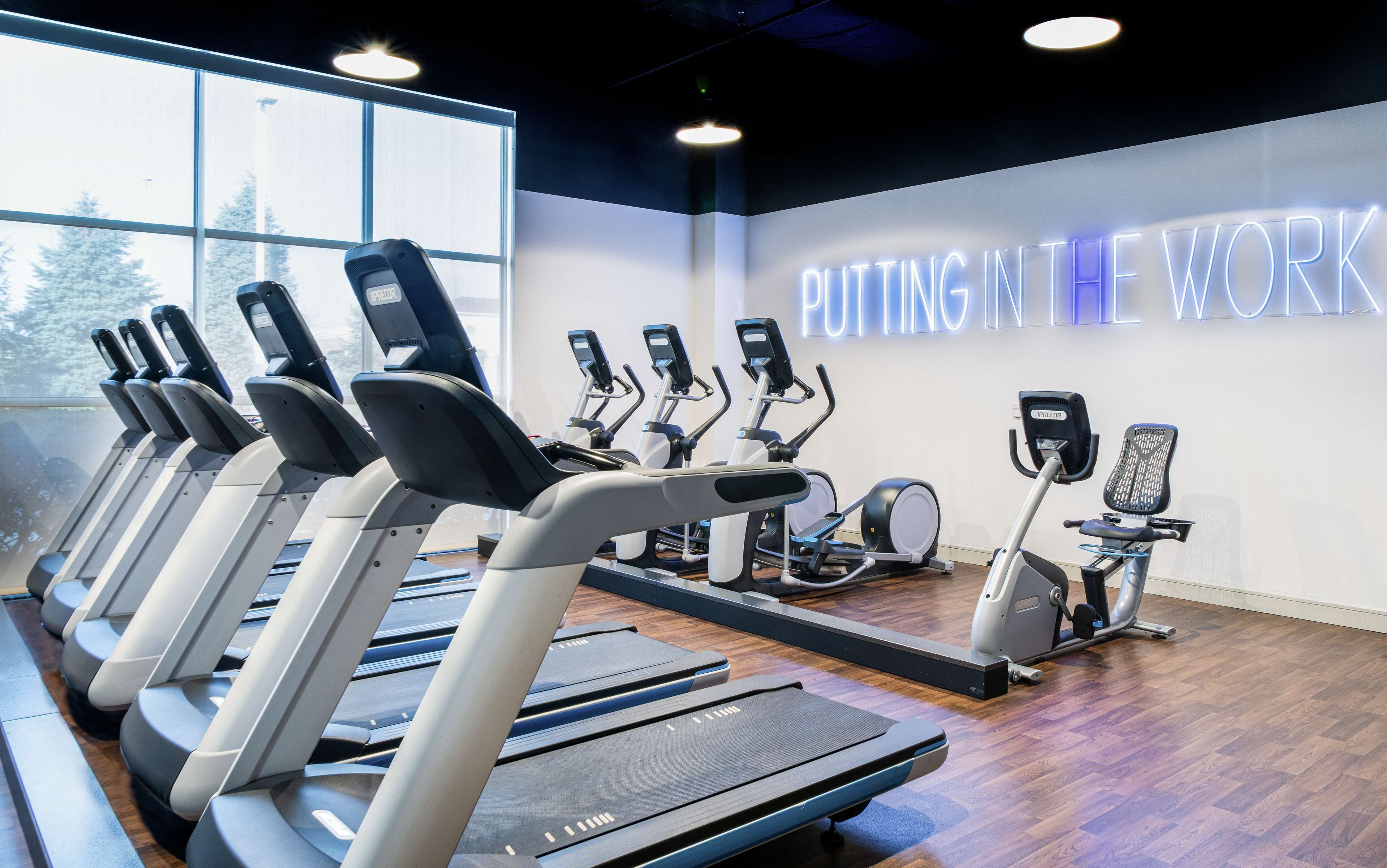 A gym with multiple treadmills, elliptical machines, and a wall sign that reads "CUTTING IN THE WORK".