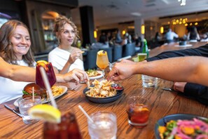 A group of people dining at a restaurant with a wooden table, plates of food, and drinks. - Hilton Columbus/Polaris (Columbus)