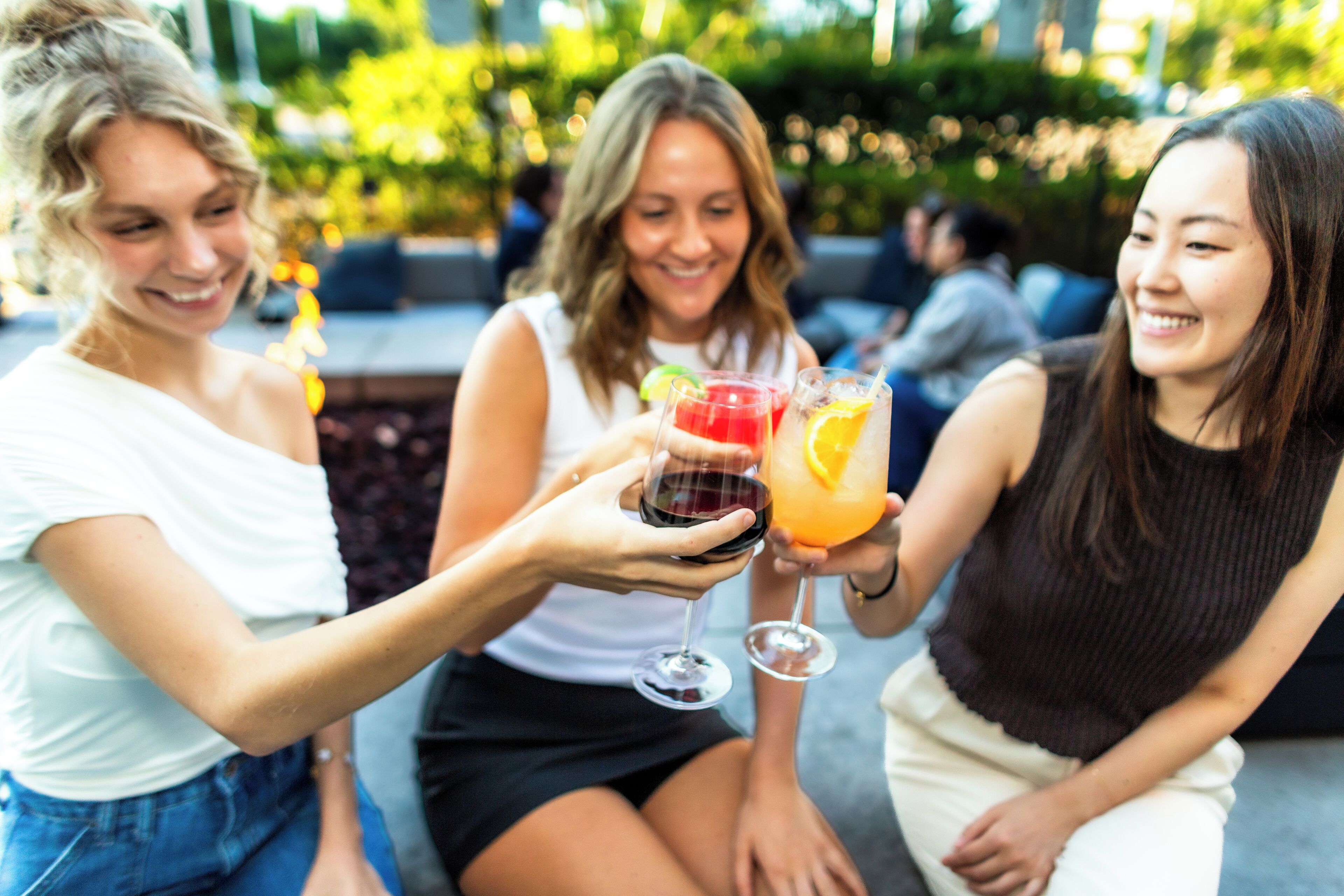 Drie personen die met drankjes proosten, buiten gezeten, met een vage achtergrond die een sociale gelegenheid suggereert.