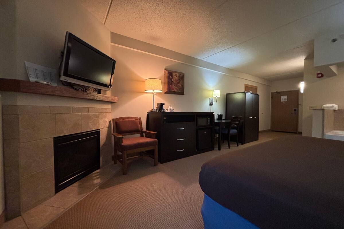 A hotel room with a flat-screen TV mounted above a fireplace, a desk with a lamp, a chair, and a bed.