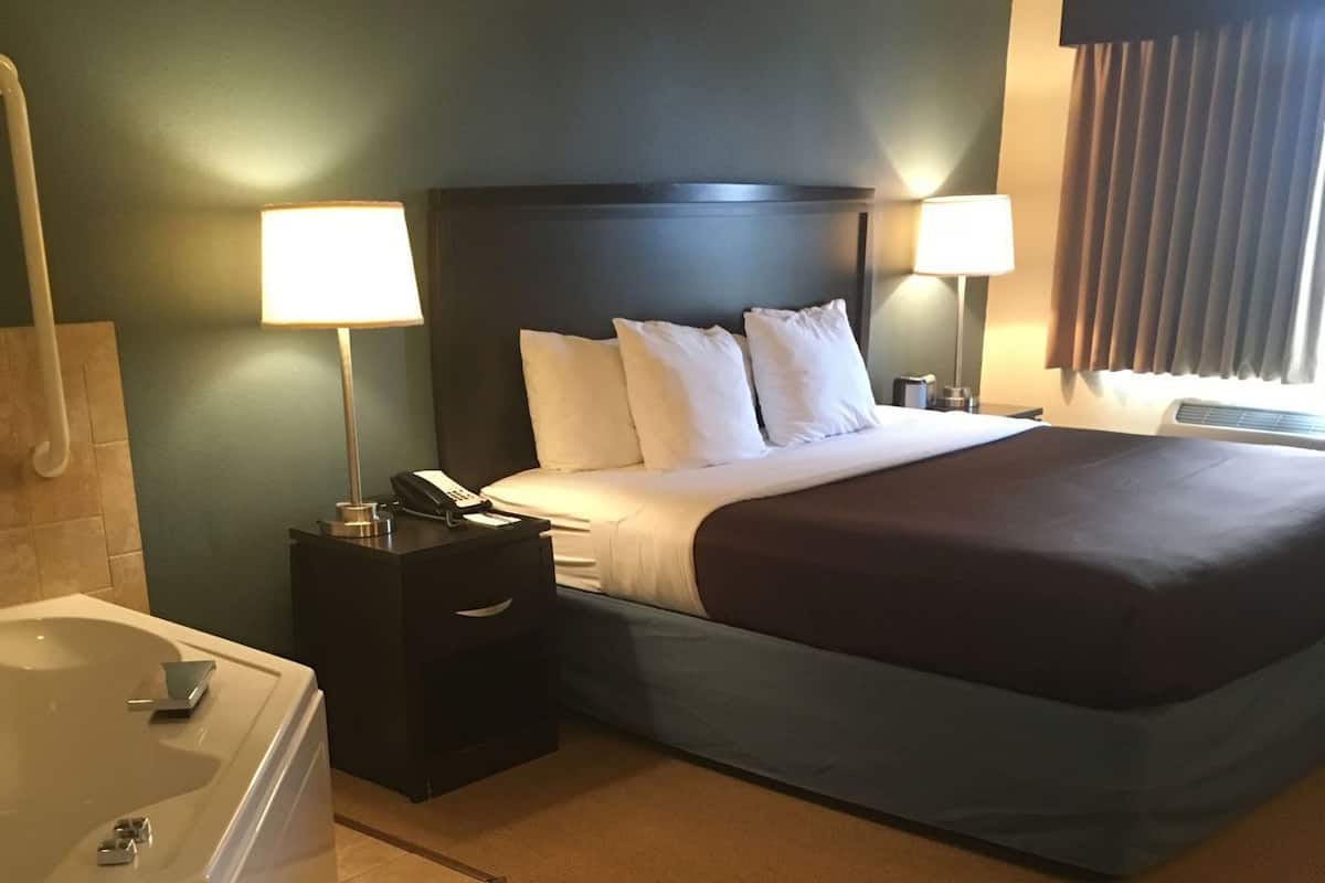 A hotel room with a large bed, two bedside lamps, a nightstand, a bathroom with a bathtub, and a window with curtains.