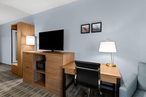A hotel room with a wooden desk, a flat-screen TV, a black office chair, a lamp, and a small sofa.