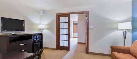 Quarto de hotel com escrivaninha, televisão, duas luminárias, um sofá e uma porta que dá para outro quarto.