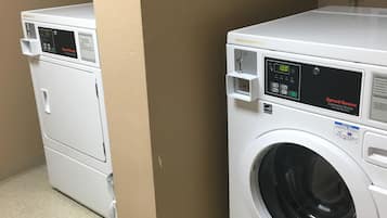 A laundry facility with two front-loading washing machines, a sign indicating where laundry supplies are sold, and a tiled floor.