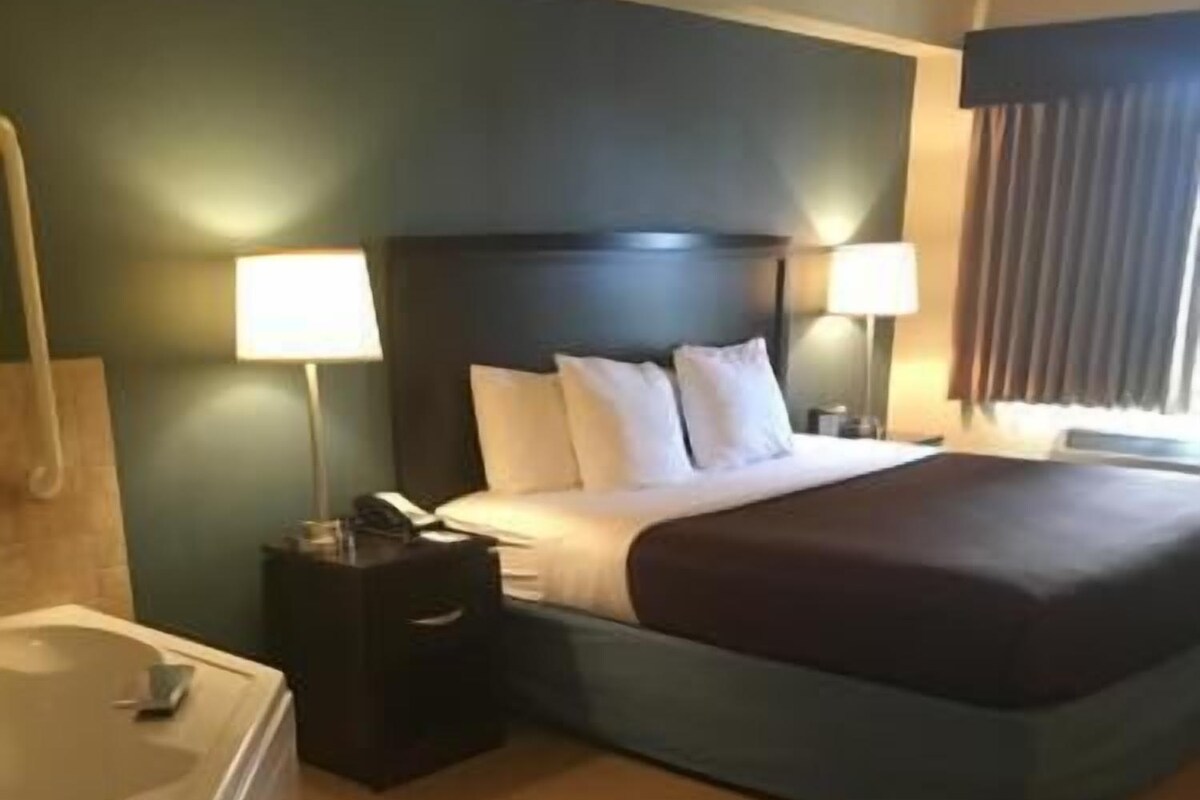 Habitación de hotel con cama, dos lámparas de noche, mesita de noche, baño con bañera y ventana con cortinas.