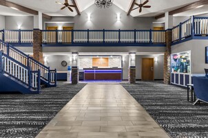 A hotel lobby with a central reception desk, a staircase, and a high ceiling with fans.