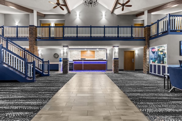 Un vestíbulo de hotel con un mostrador de recepción central, una escalera y un techo alto con ventiladores.