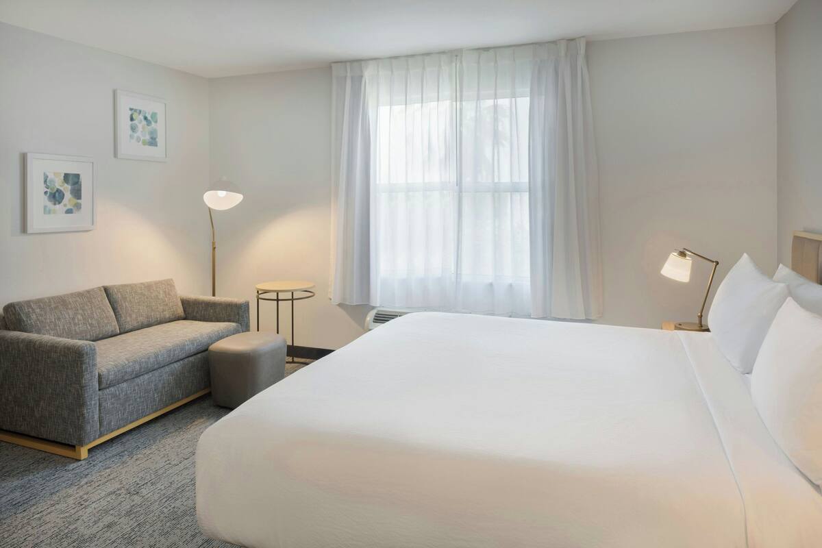 Ett hotellrum med en stor säng, en grå soffa, ett litet bord och två väggmonterade lampor.